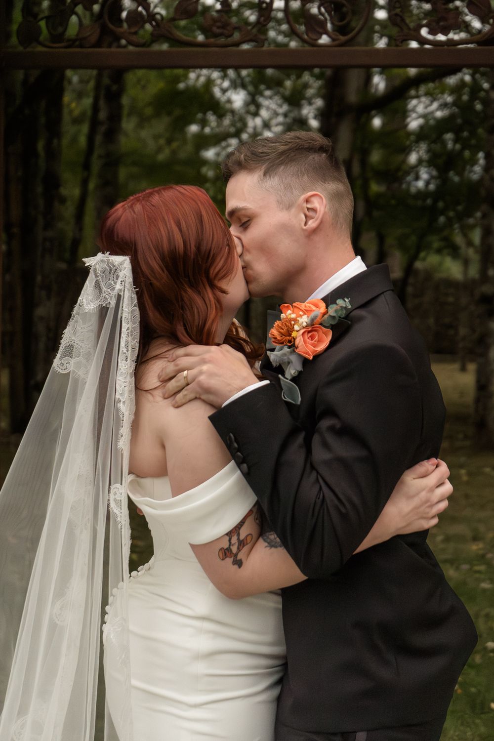 A romantic wedding moment captured in a wooded setting with the couple sharing a kiss, featuring orange floral accents.