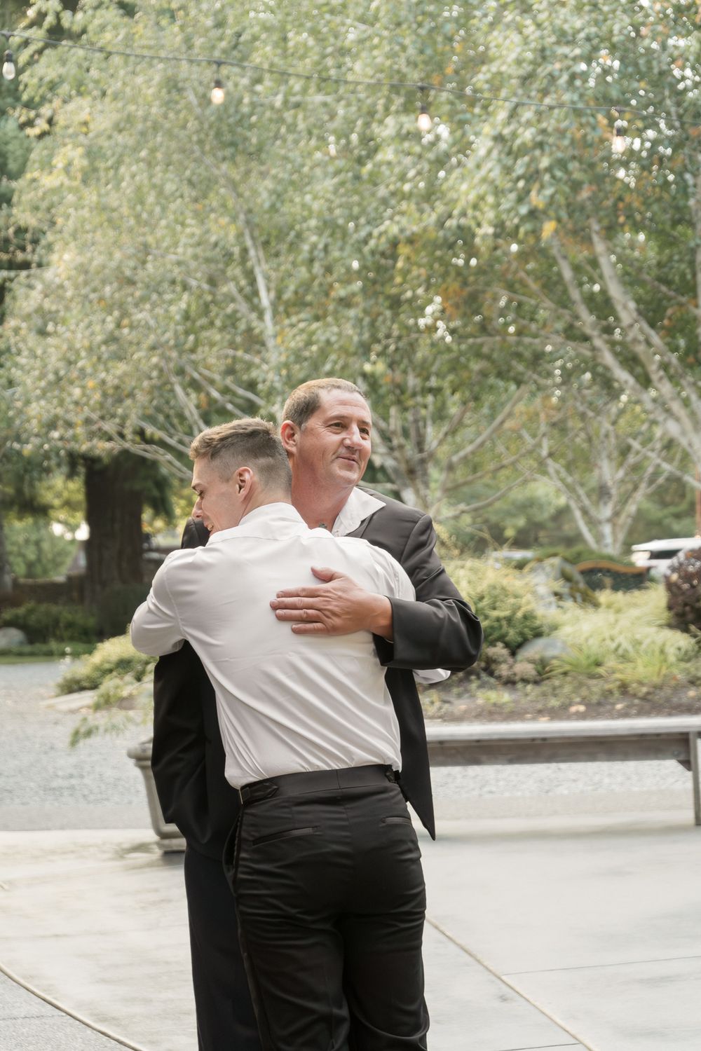 Two people in formal attire share an emotional embrace outdoors on a tree-lined path.