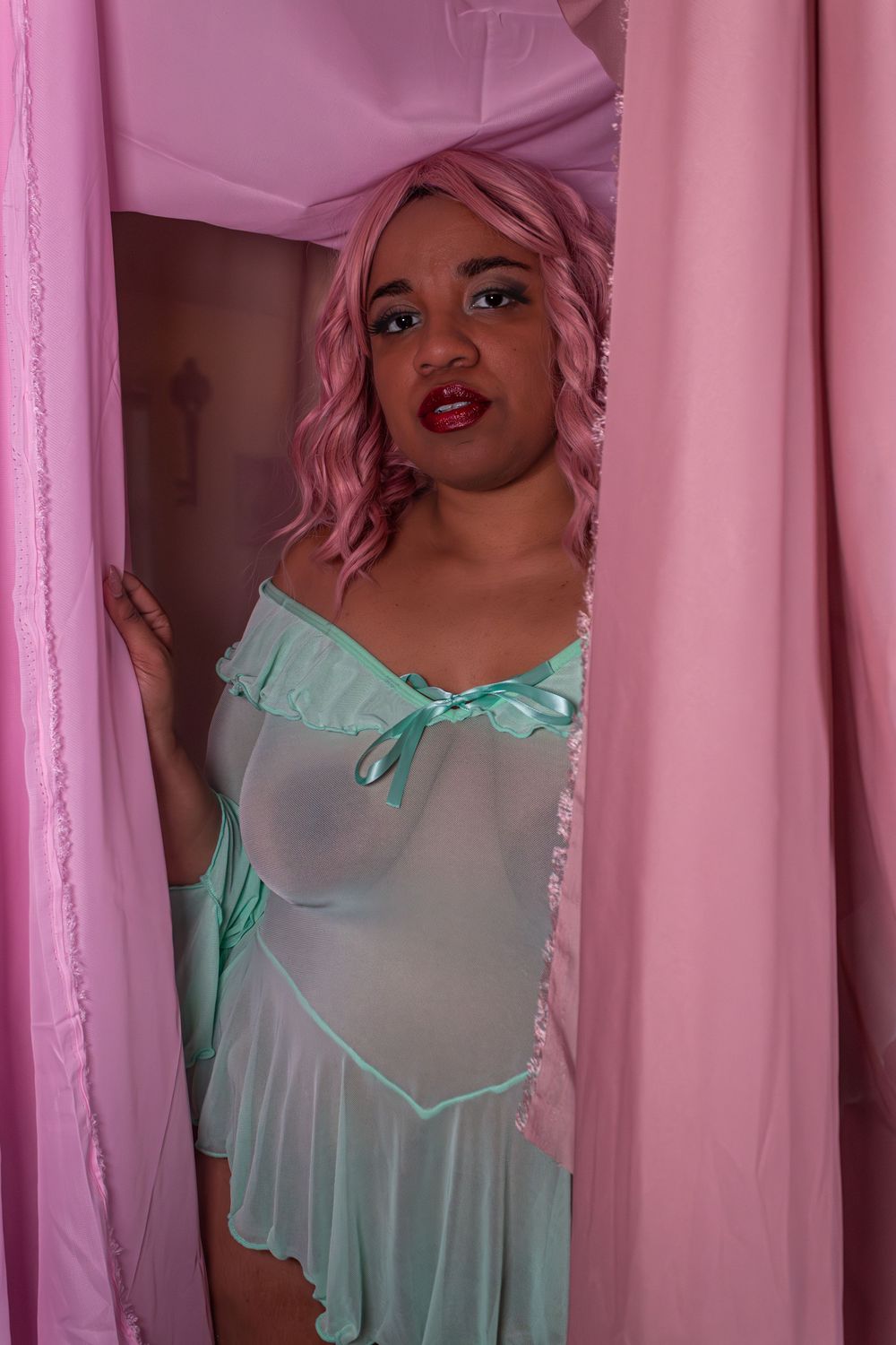 Woman with pink hair in mint green sheer dress poses between pink curtains.