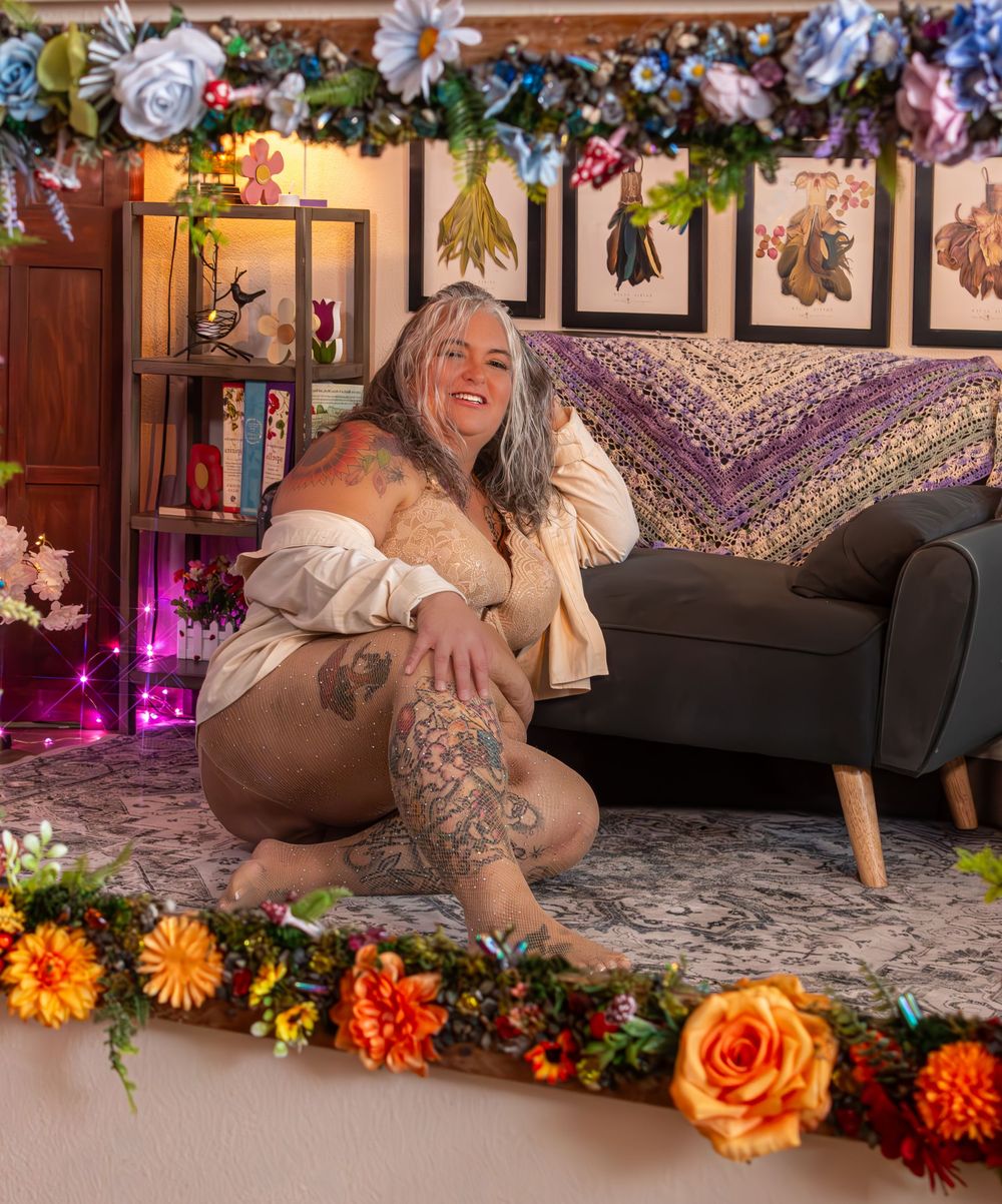 Tattooed woman with gray hair sits smiling on a floral-decorated floor beside a gray sofa in a cozy, art-filled room.