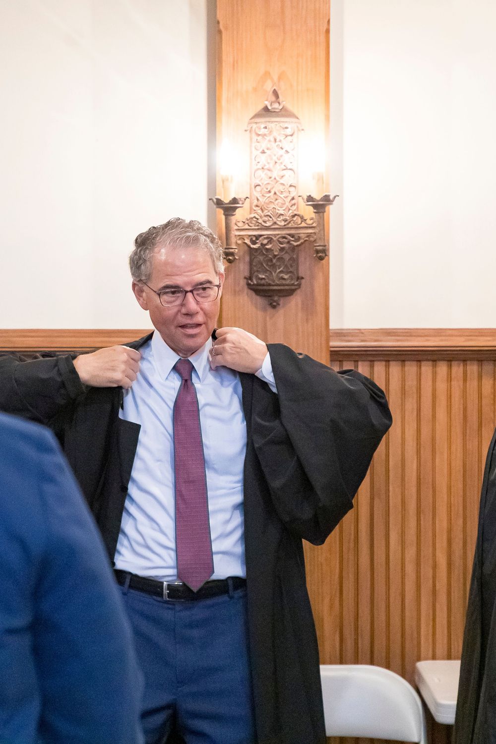 Associate Justice Mendheim in judicial robes adjusting his robe in a wood-paneled room.