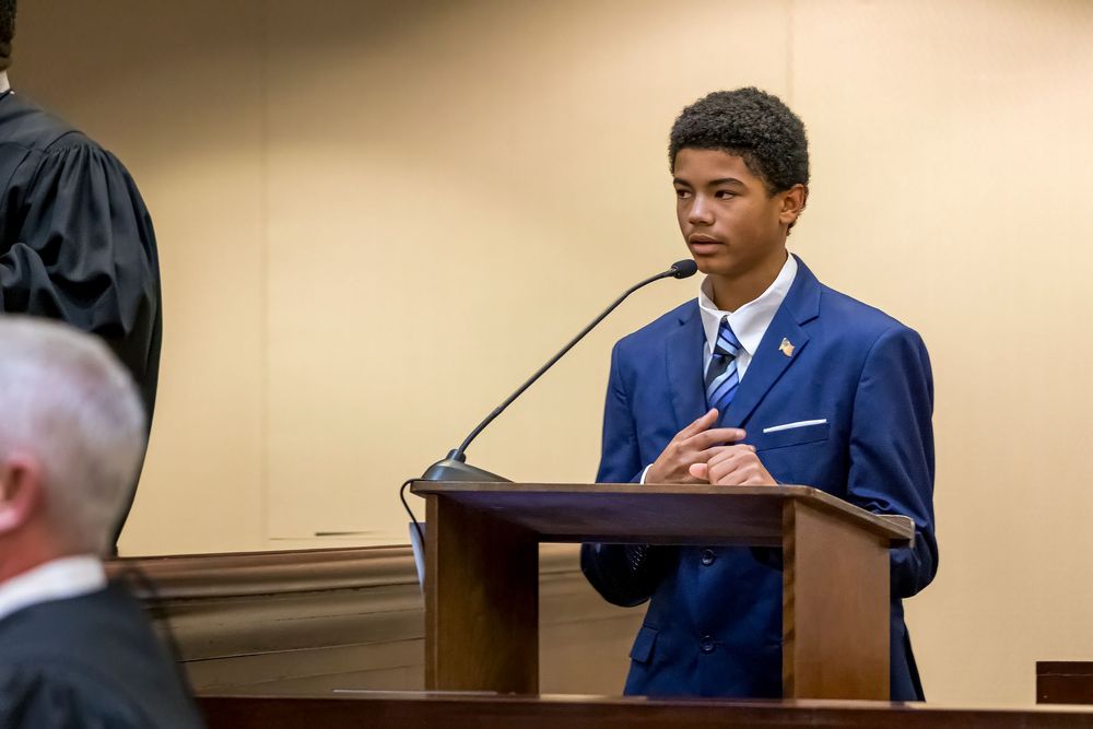 A person in a navy suit sits at a desk during what appears to be a formal hearing or testimony.