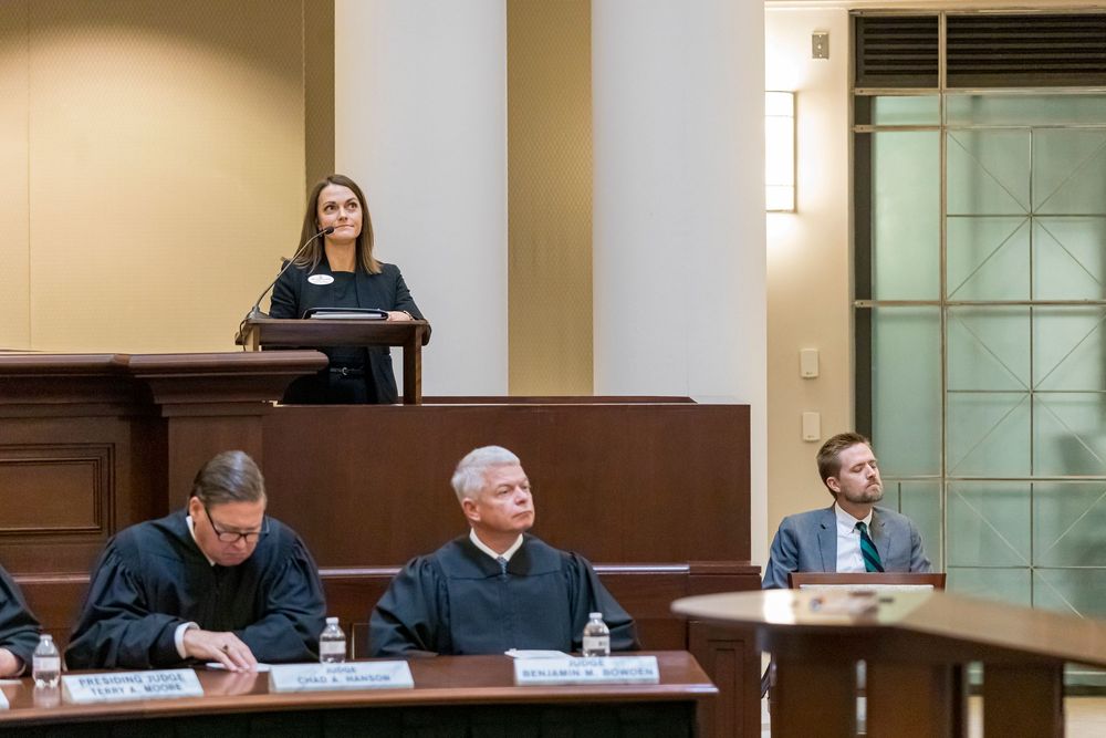 Court proceedings in session with officials seated at bench while presenter speaks from podium.