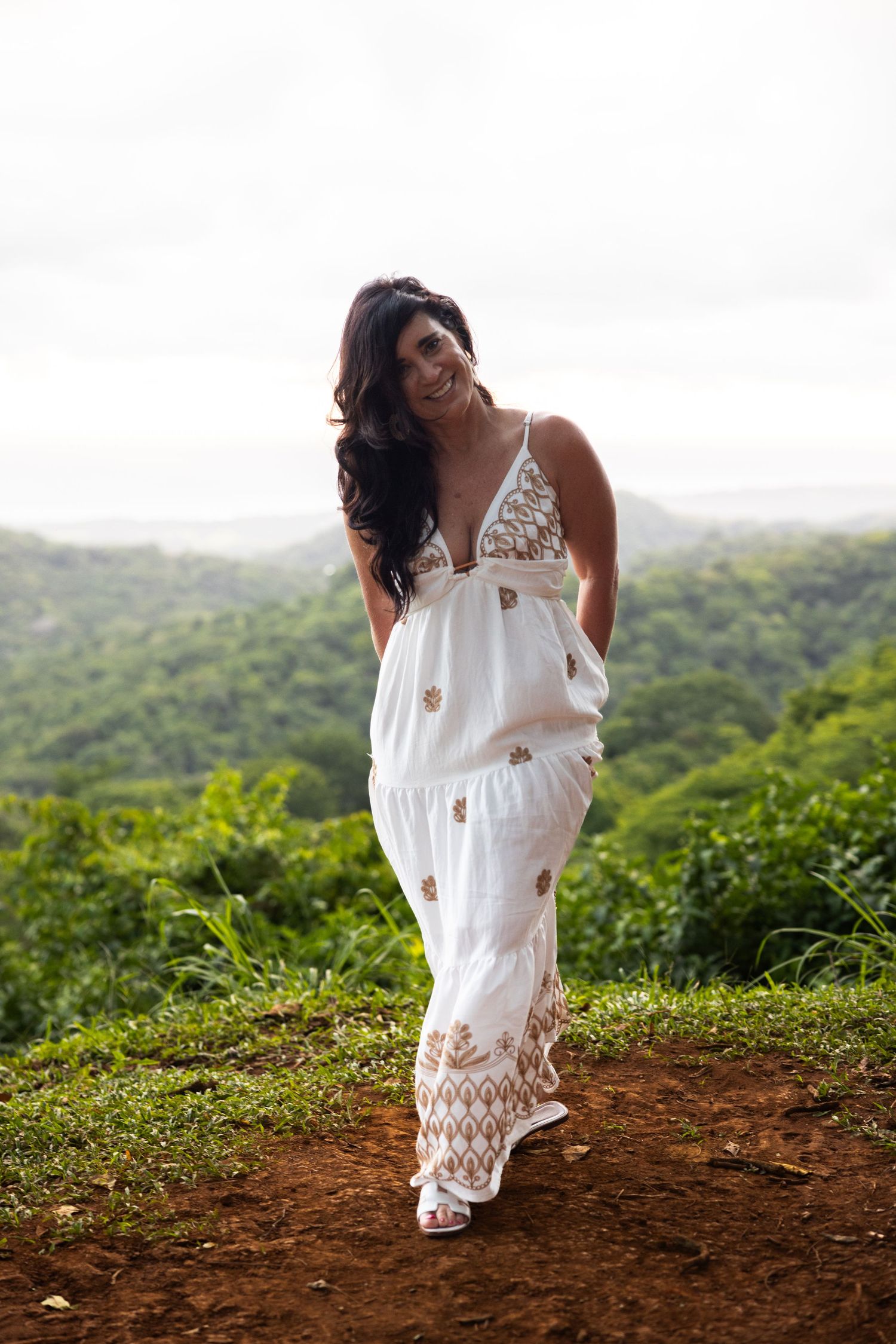 Person in flowing white dress walking along dirt path with tropical hillside views.