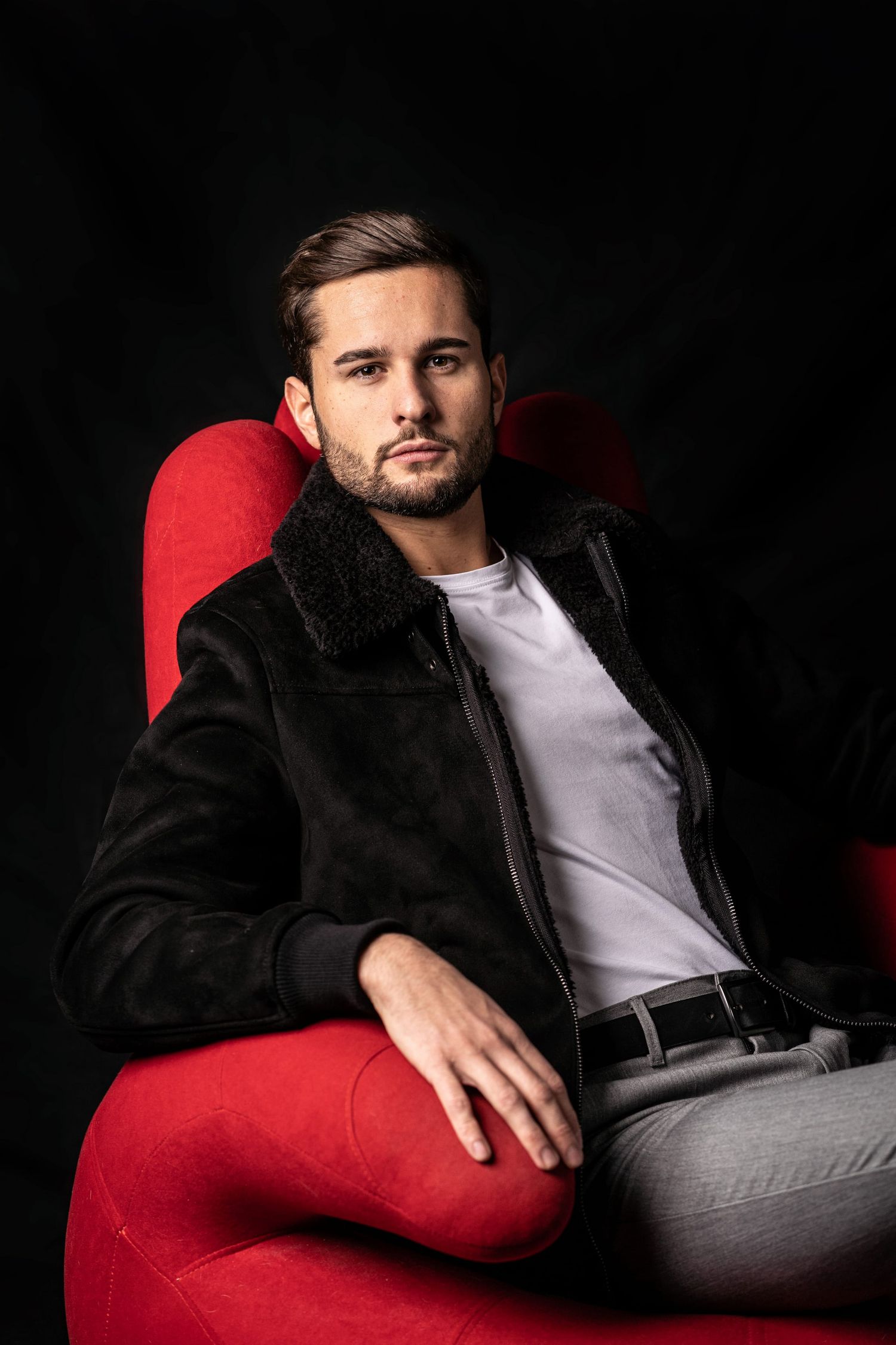 Stylish man in black shearling jacket sitting in red armchair against dark studio background.