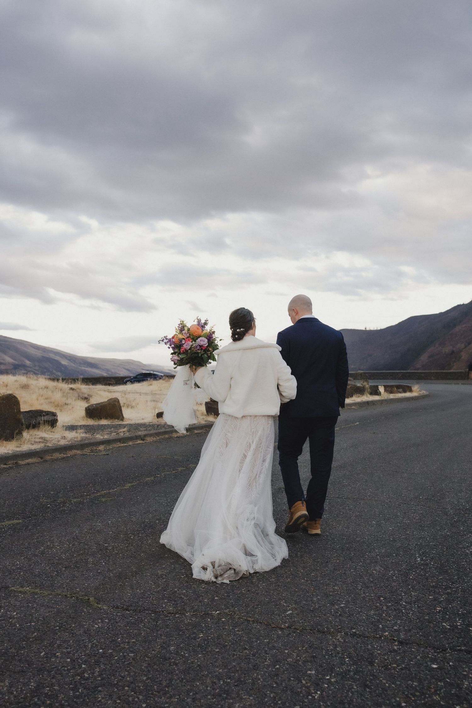 A romantic wedding couple embraces on a stone wall against a dramatic cloudy mountain landscape.