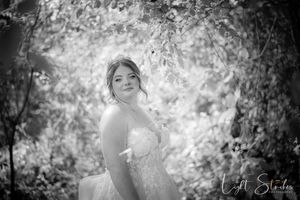 A black and white art edited photo shows a bride in a white dress posing gracefully among trees and foliage.