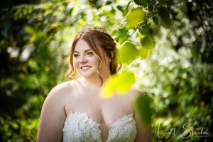 A bride in a white lace wedding dress smiles radiantly in a sun-dappled garden setting with green foliage.