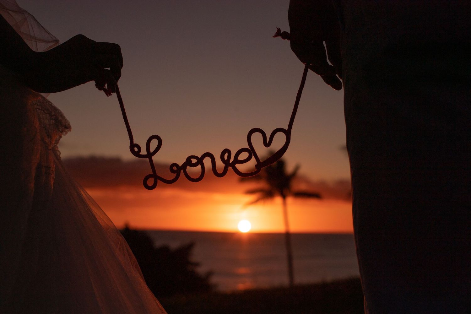 Couple holding a wire Love sign at sunset on a beach, with a palm tree and ocean in the background. サンセットフォトウェディング