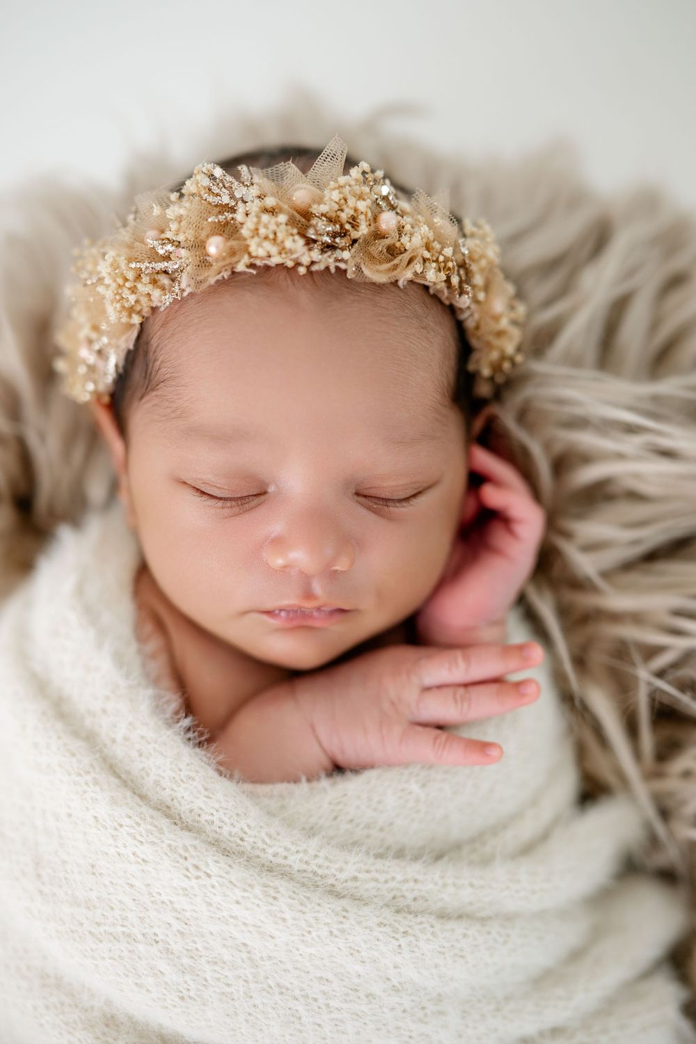 Sleeping newborn wrapped in soft white blanket wearing delicate floral crown in dreamy studio portrait series.