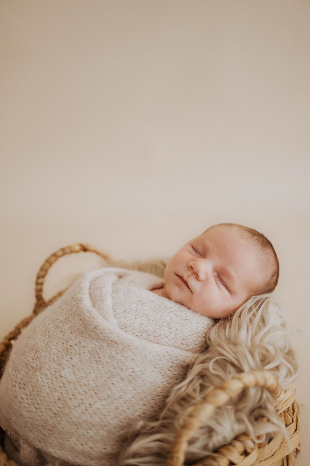 Sleeping baby wrapped in soft white knit blanket lying on textured beige surface.