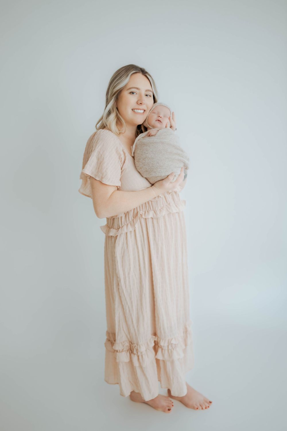 A parent and baby wearing matching soft pink dresses share tender moments against a light gray backdrop.