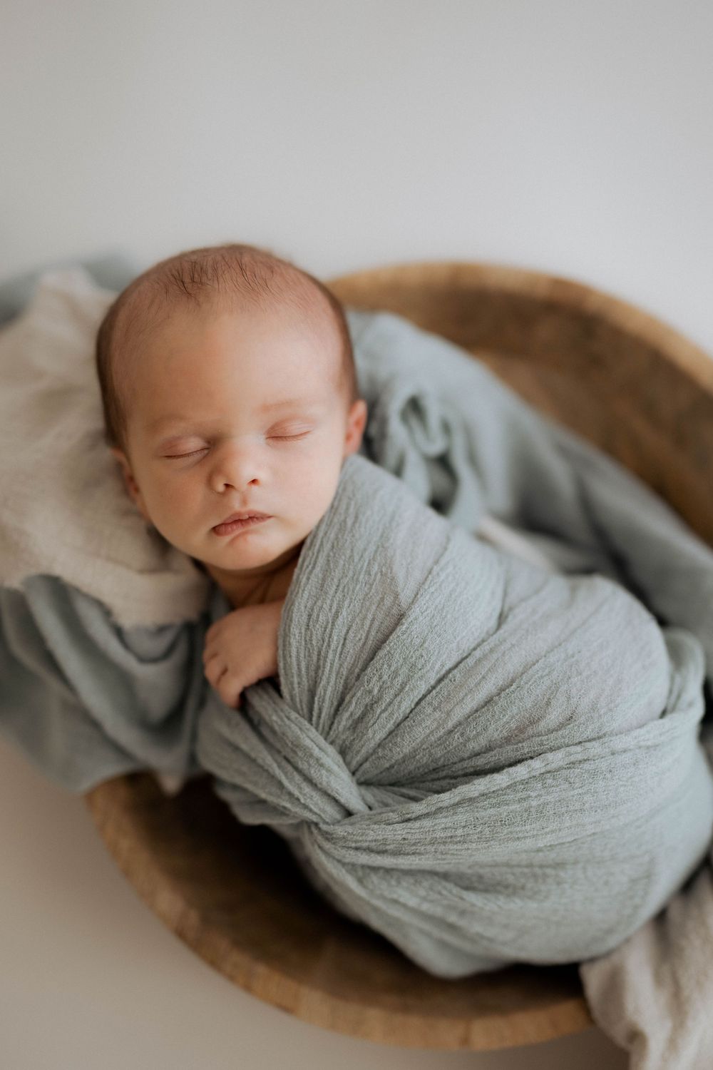 Sleeping newborn baby wrapped in soft sage green swaddle blanket resting in wooden bowl during professional photoshoot.