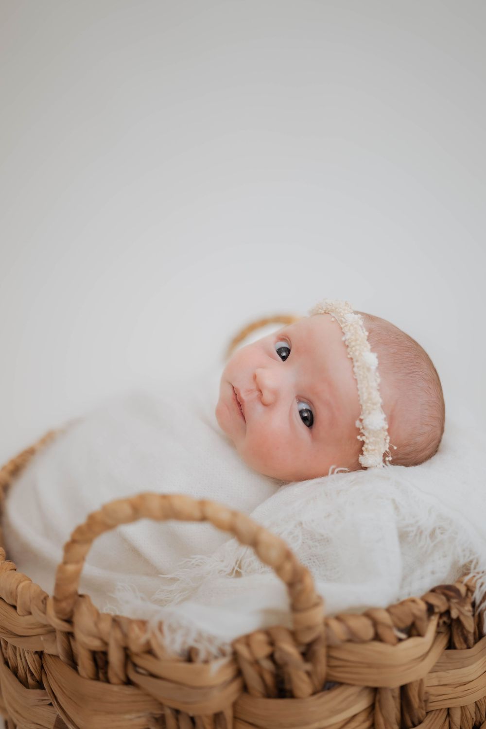 A baby wrapped in white fabric lies in a woven basket against a light gray background.