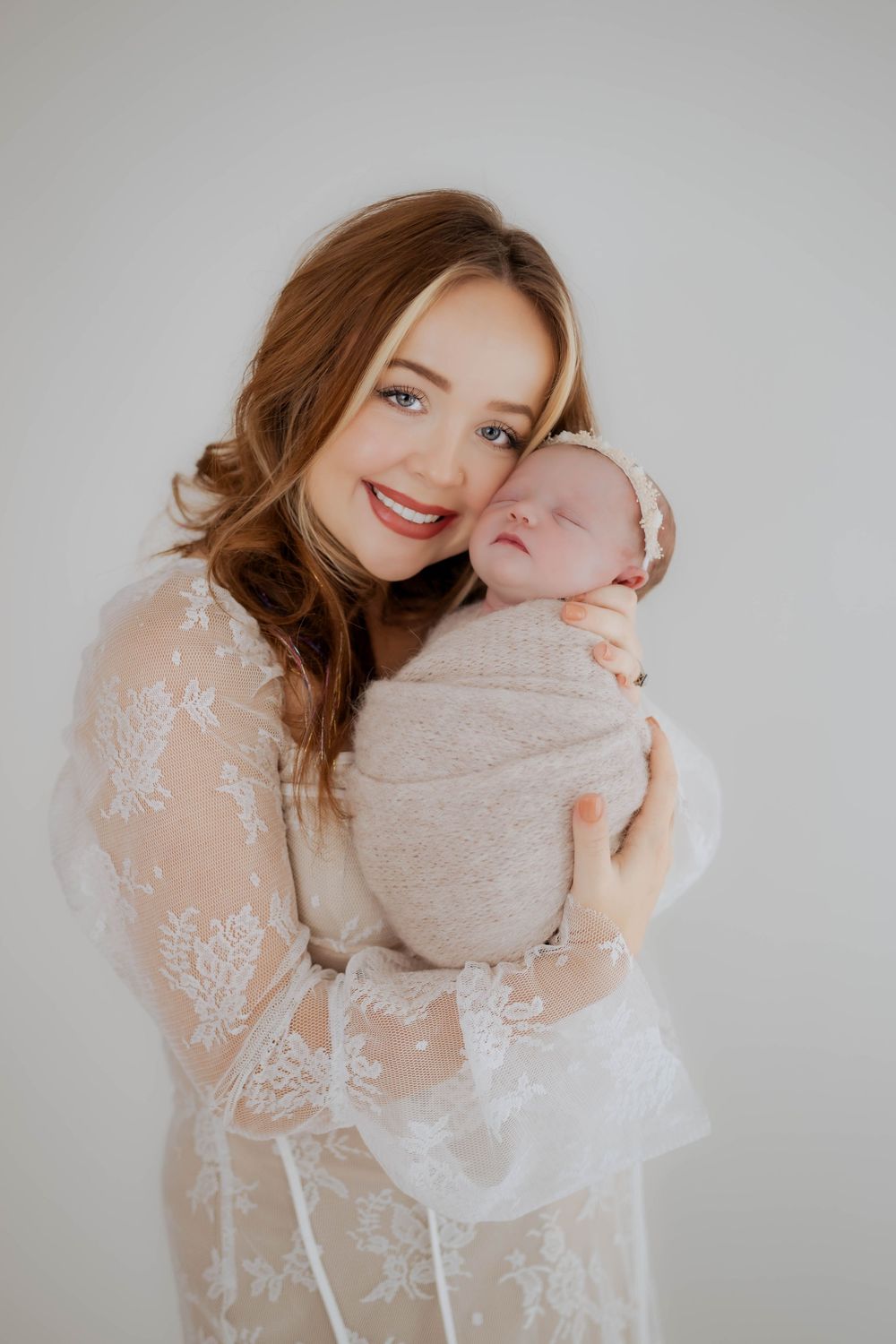 Parent in delicate lace dress embracing sleeping newborn against light backdrop.