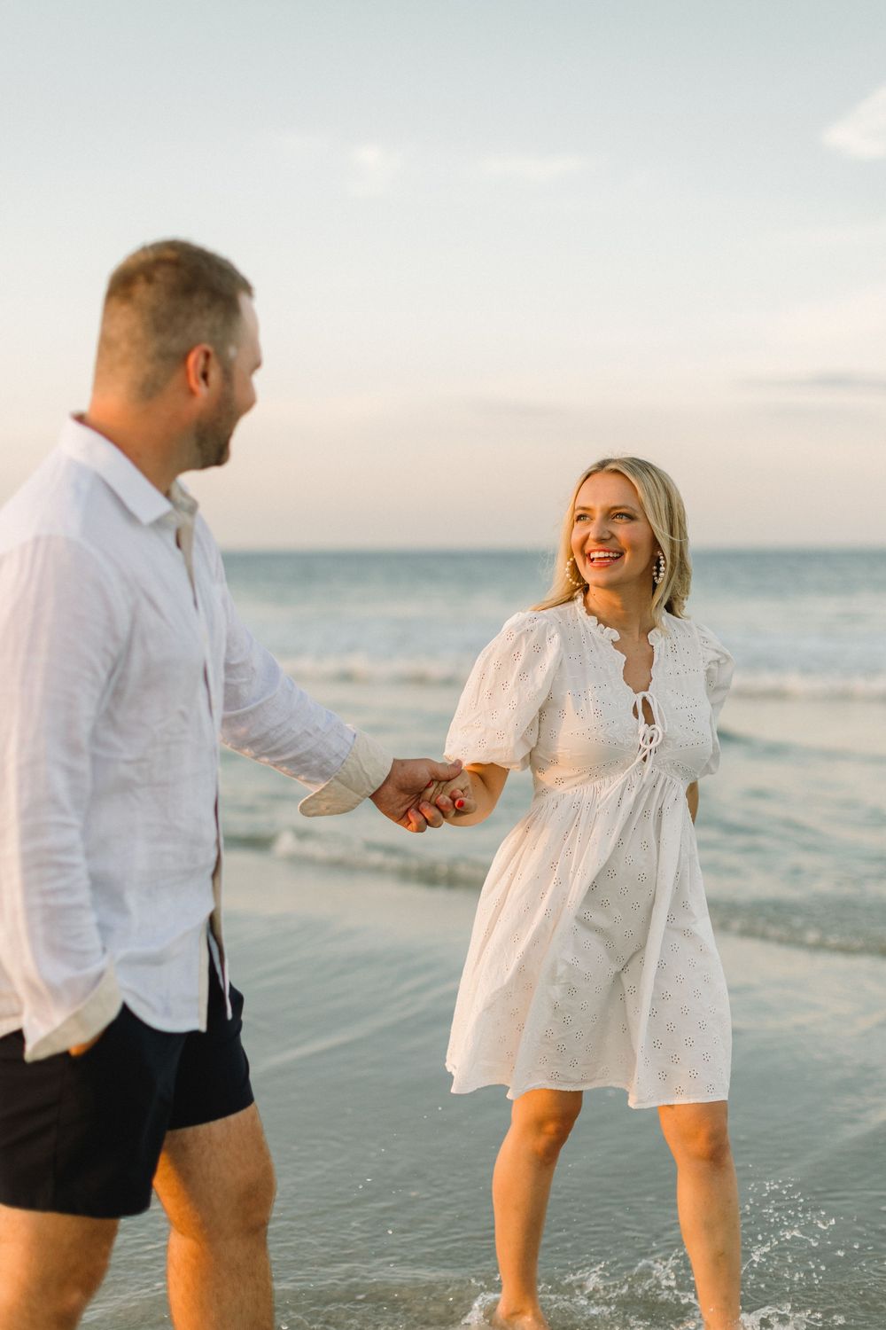 A couple in white clothing shares a playful moment together on a sunny beach at sunset.