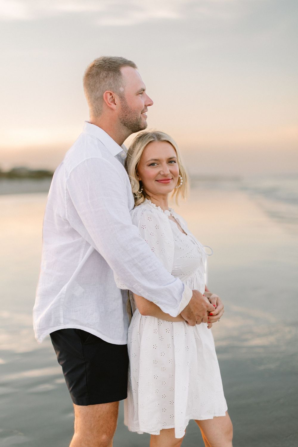 A couple in white clothing shares romantic moments on a beach at sunset.
