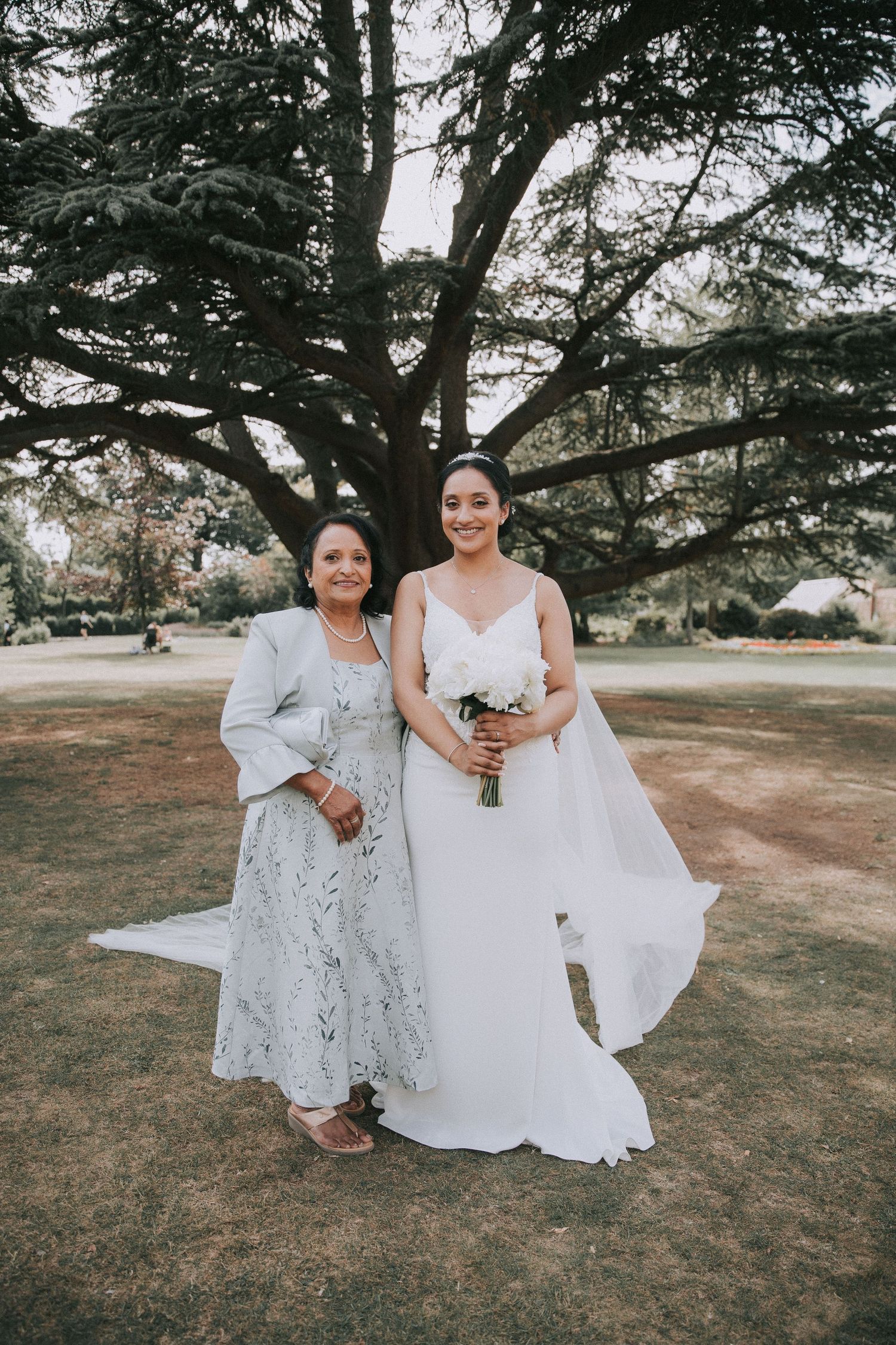 Two people in formal attire pose together on a grassy lawn with trees in the background during a wedding photoshoot.