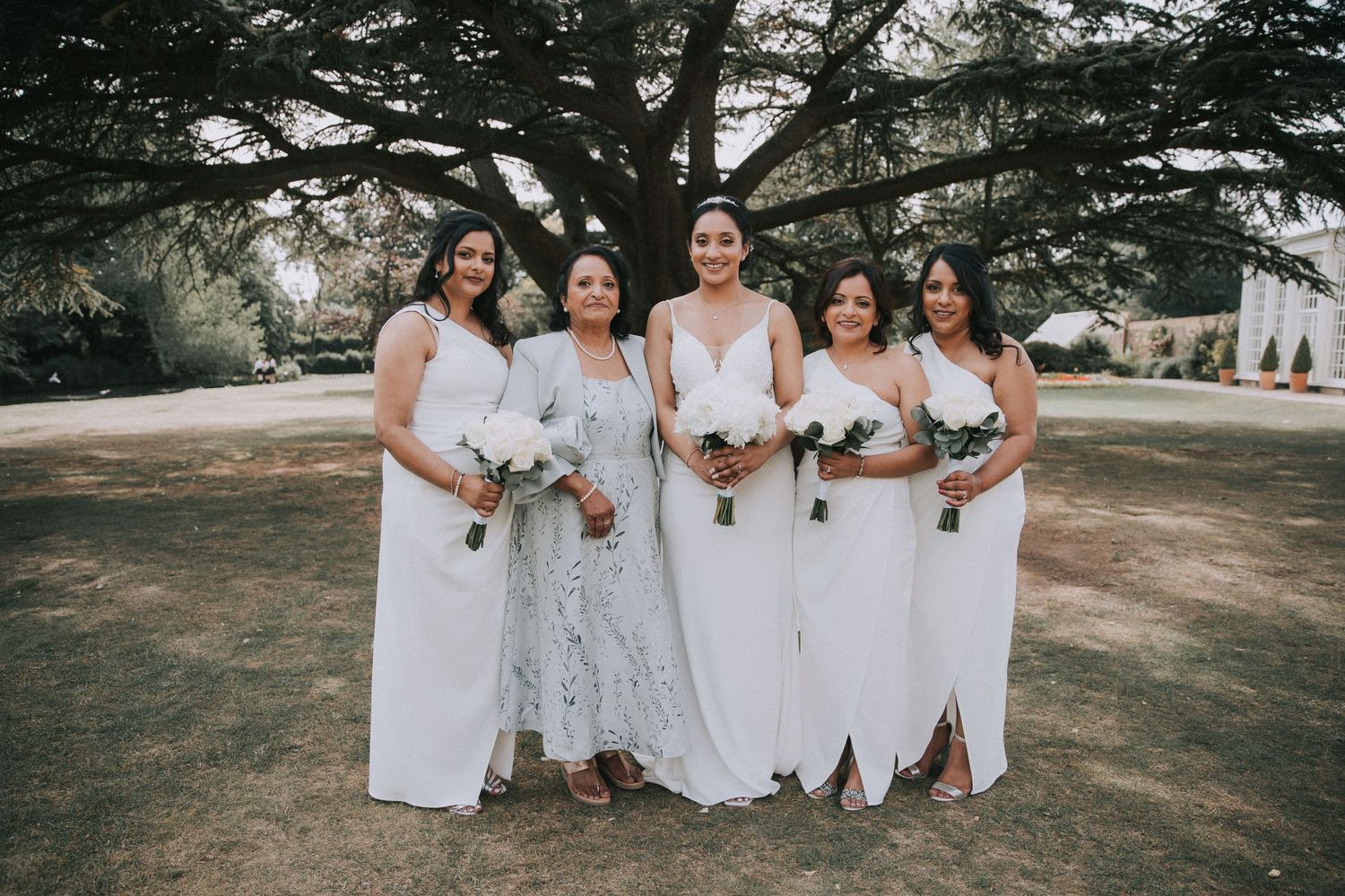 A wedding party in white dresses poses together on a grassy lawn surrounded by tall trees.