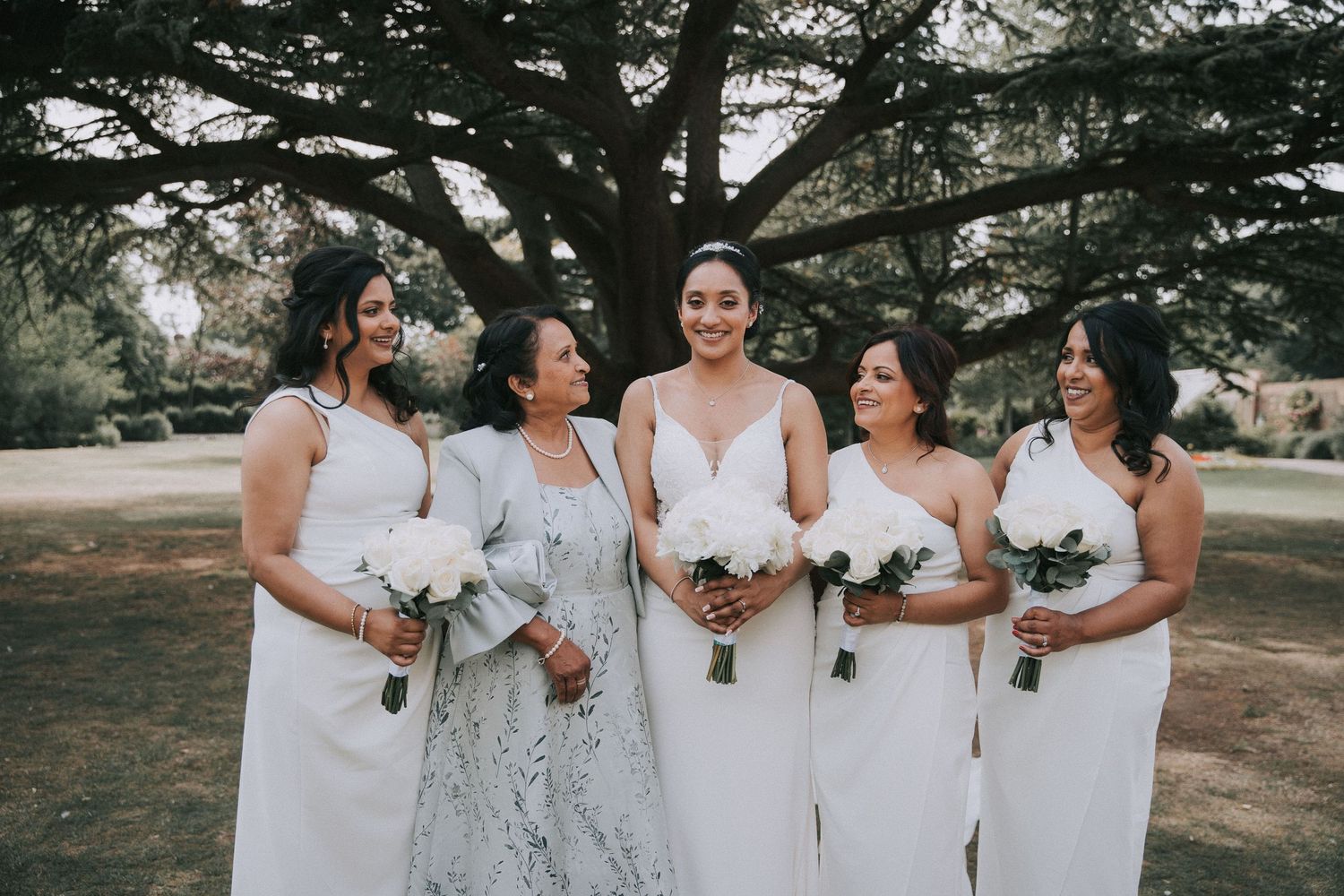 A group in white dresses poses together outdoors under large trees, creating an elegant wedding party scene.