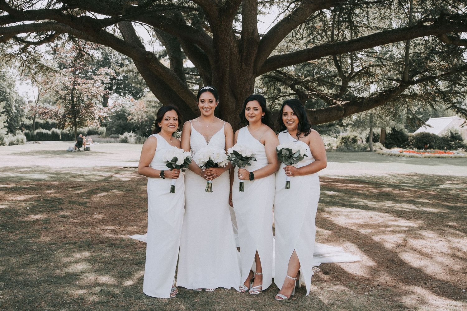 Wedding party members in white dresses stand together under shady trees in a natural outdoor setting.