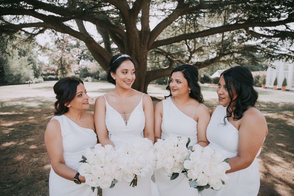 Bridesmaids in white dresses share joyful moments together under trees at an outdoor wedding celebration.