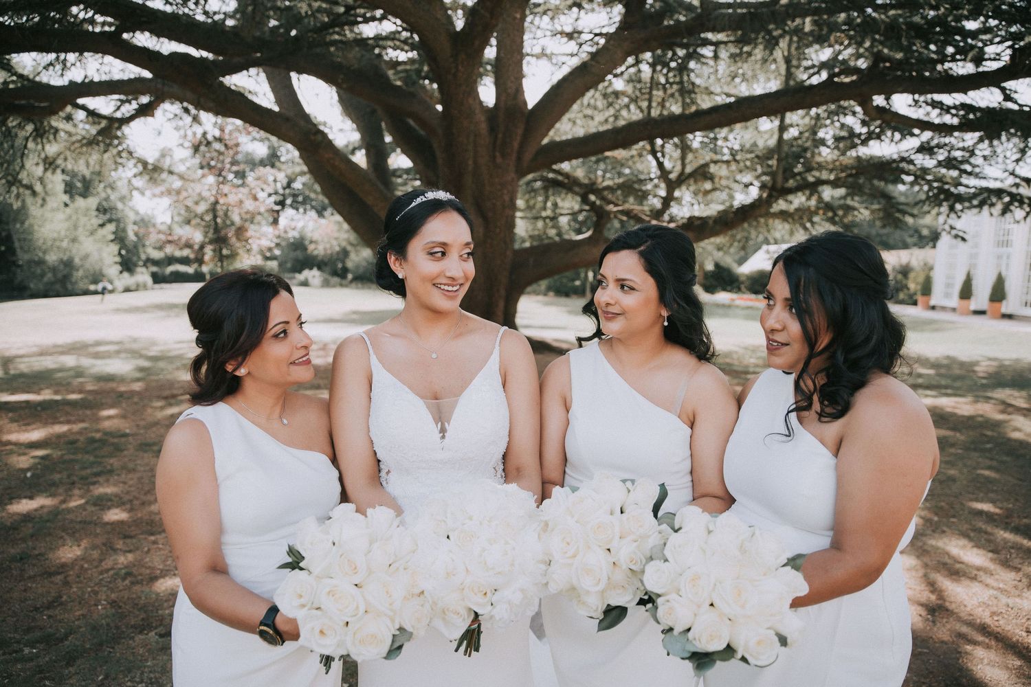 Bridesmaids in white dresses share joyful moments together under trees at an outdoor wedding celebration.
