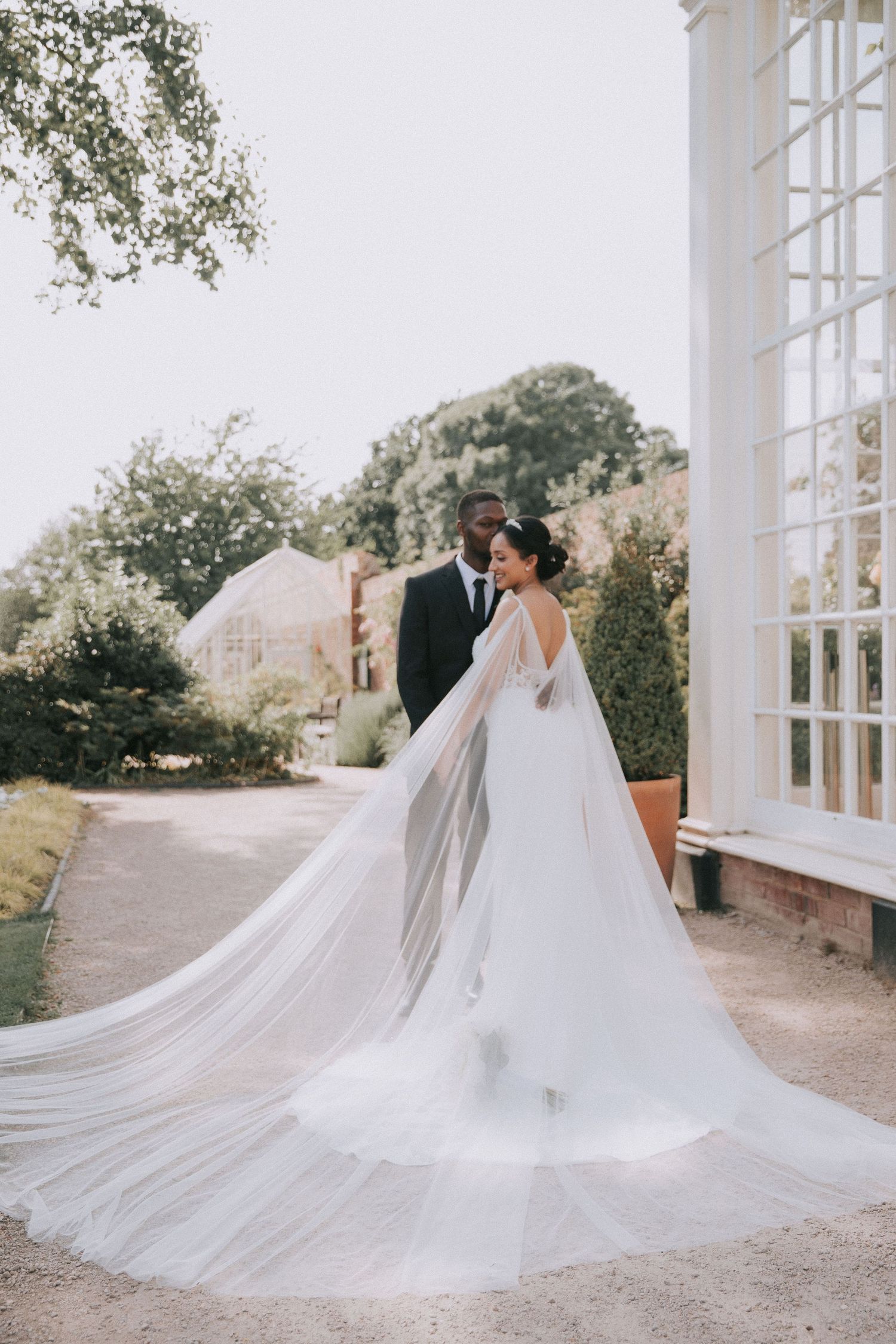 A flowing white wedding dress with a long cathedral veil photographed against a white architectural backdrop.
