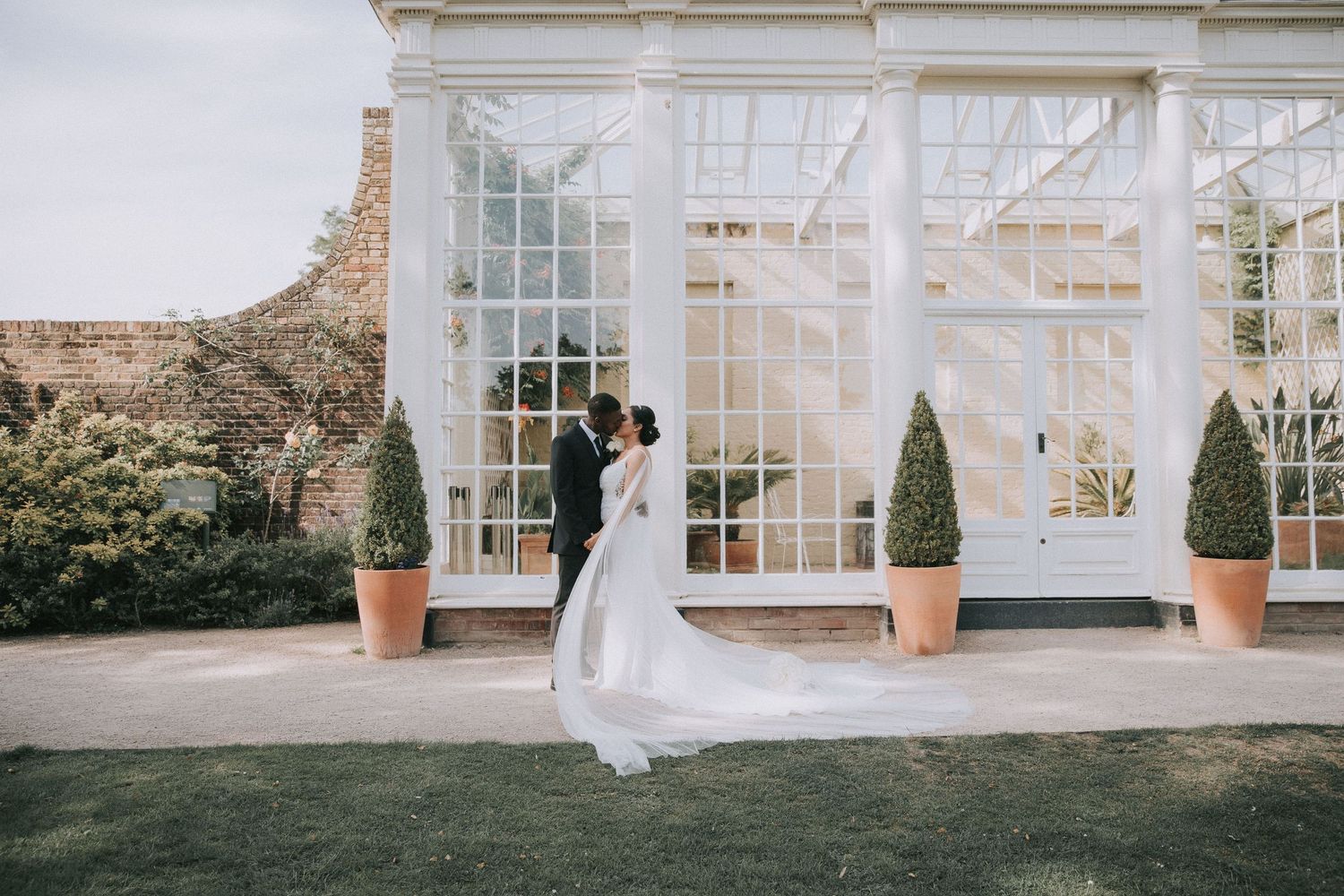 A white conservatory-style building with large windows and potted plants creates an elegant wedding venue backdrop.