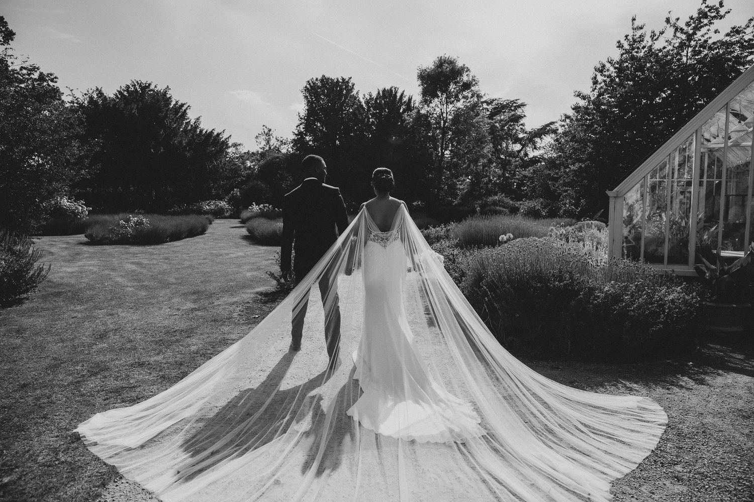 A bride and groom hold up a flowing white wedding veil in a garden setting during a photo session.