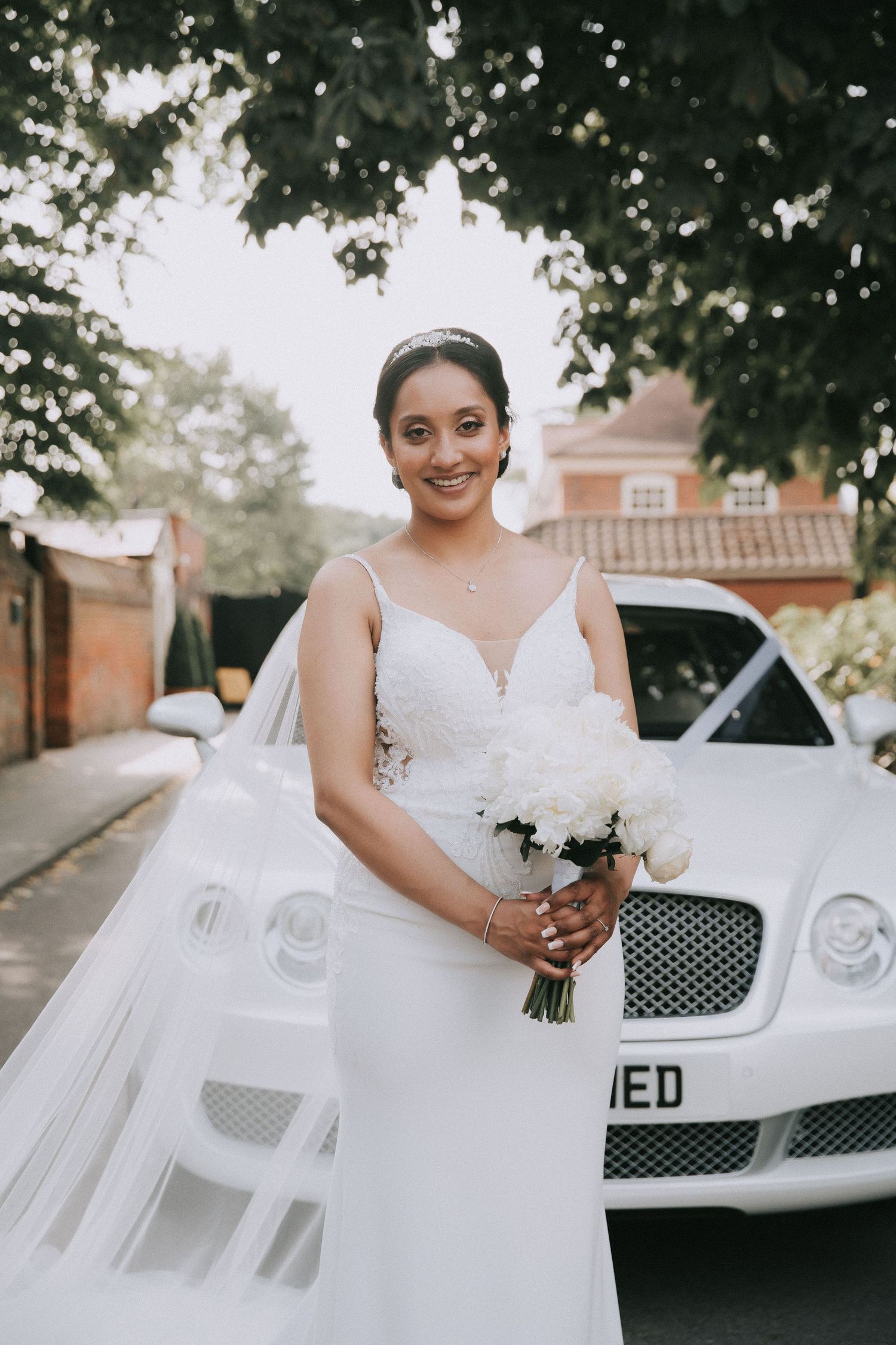 Elegant bride in white wedding dress poses next to luxury white Bentley car.