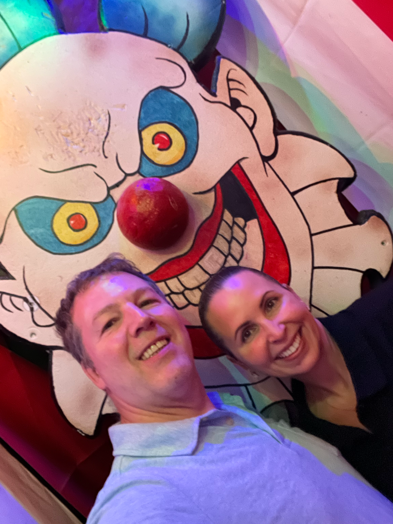 Two people take a selfie in front of a large colorful clown face decoration with glowing yellow eyes and red nose.