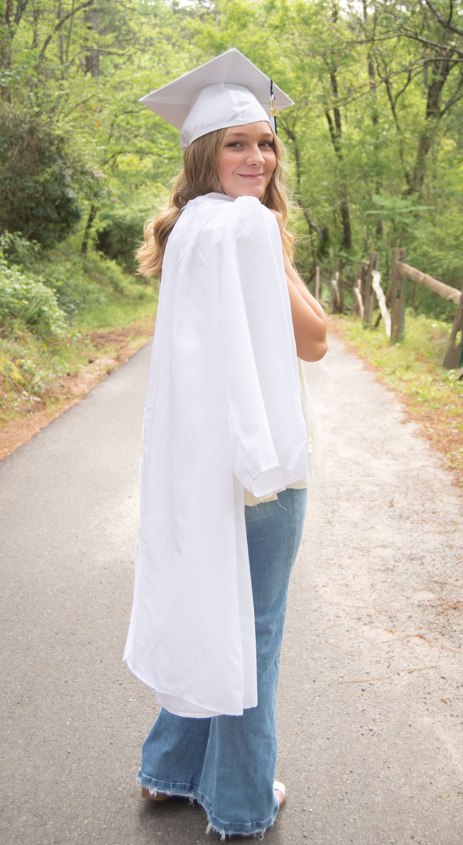 Graduate in white flowing top and jeans walks playfully down rural path.