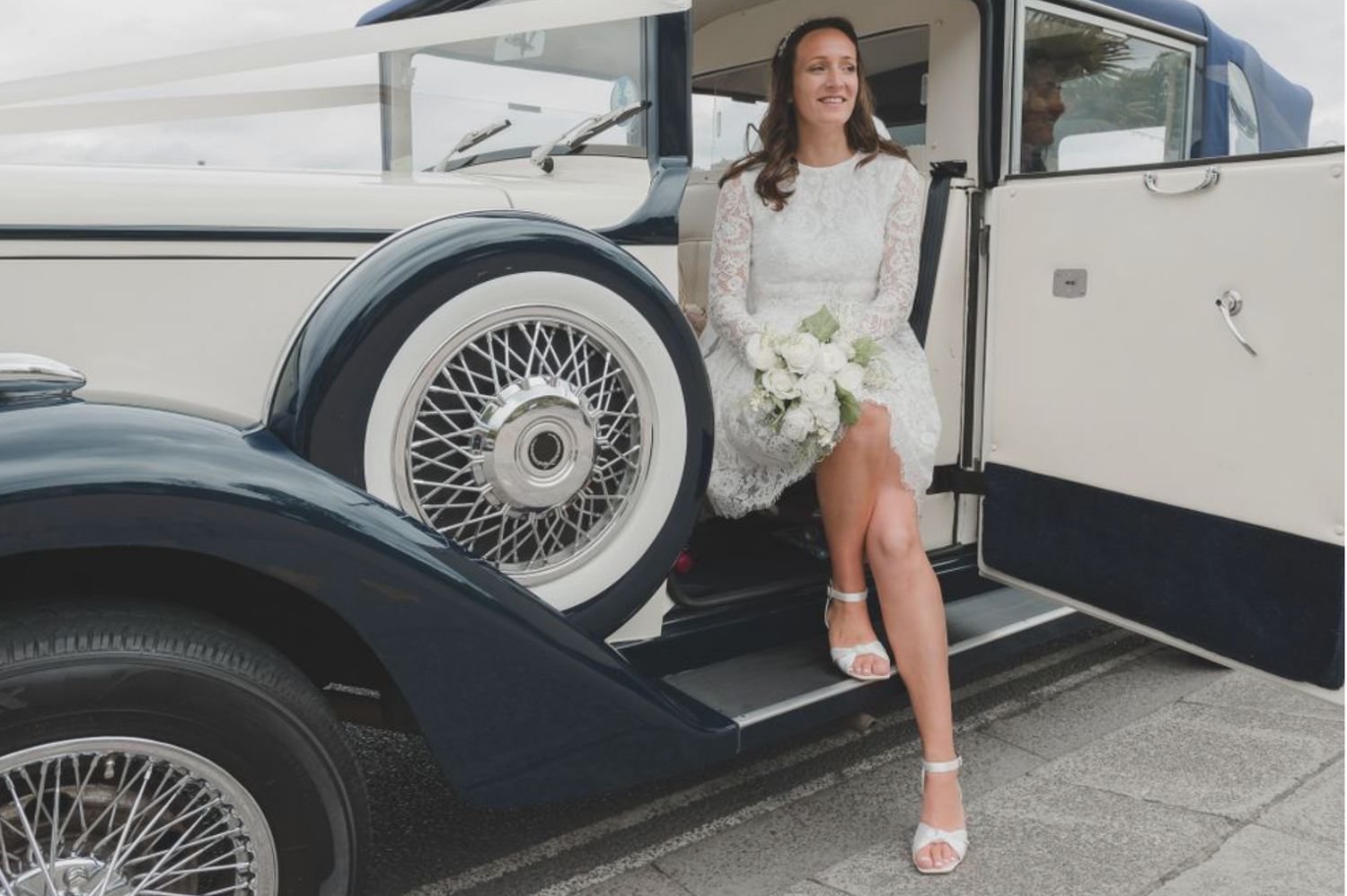 A person poses next to a classic car with vintage wire wheels and white-wall tires in a parking lot.