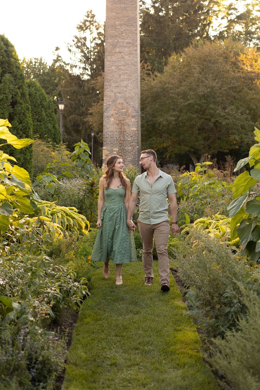 A couple walks along a garden path surrounded by lush greenery and flowers with a tall stone column in the background.