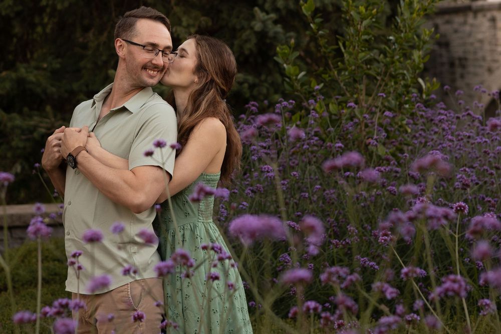 A couple embraces in a garden with vibrant purple flowers blooming in the background during a romantic photoshoot.