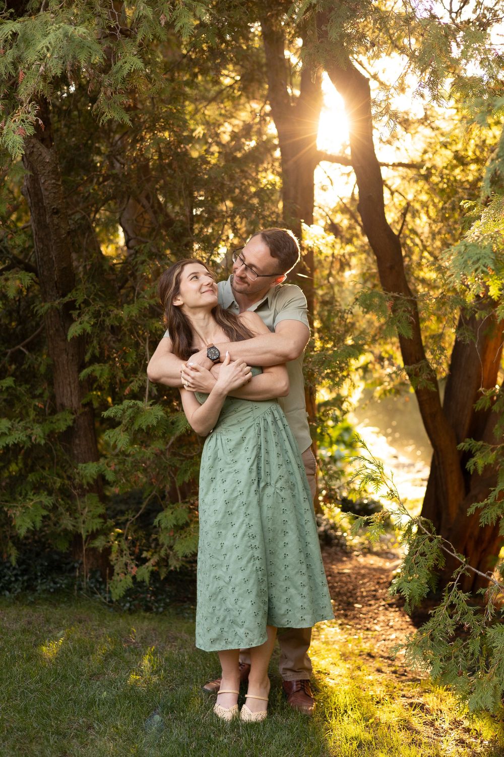 A couple embraces in a garden at sunset with golden sunlight streaming through trees in the background.
