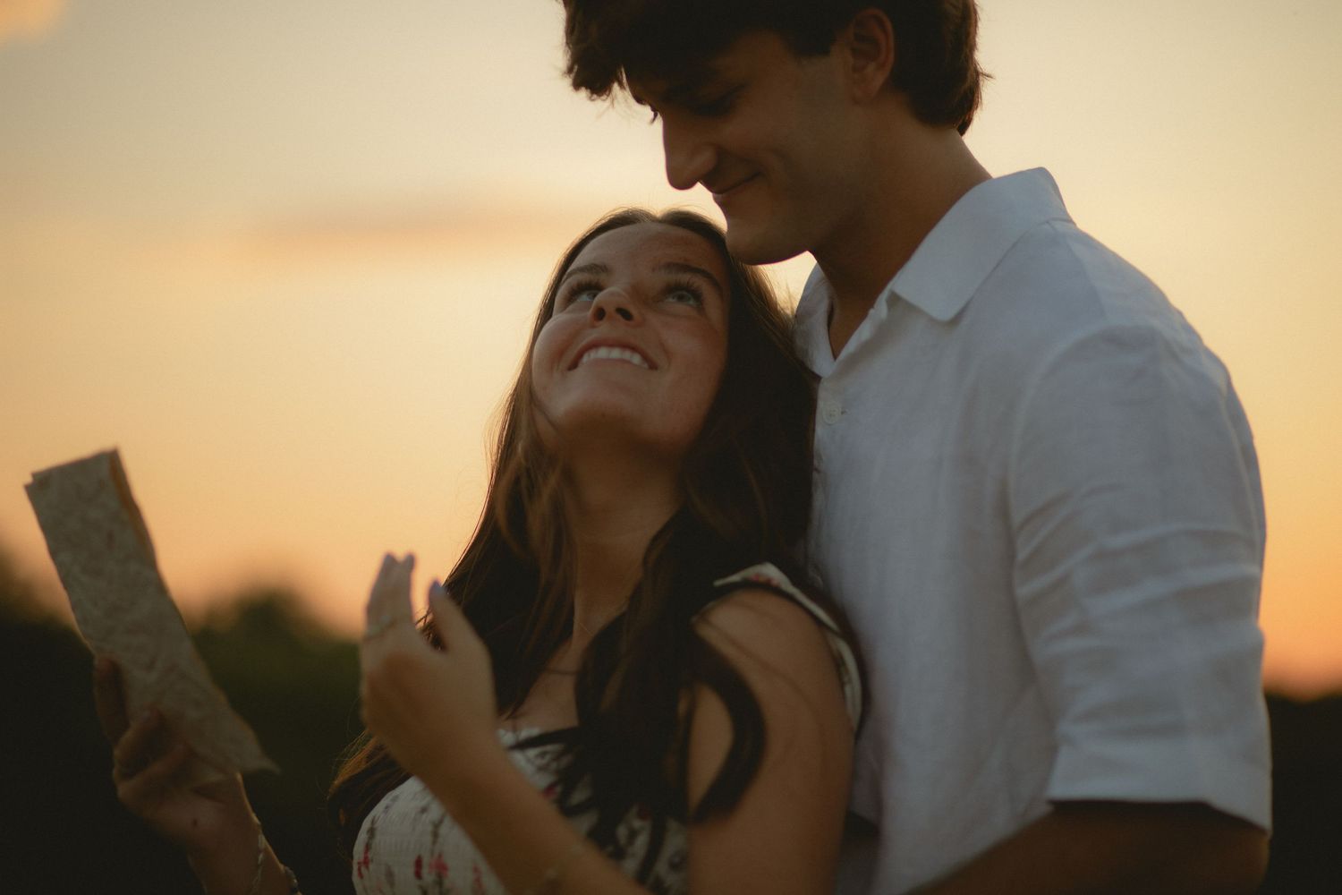 Young couple shares a romantic moment at sunset, smiling and holding a map outdoors.