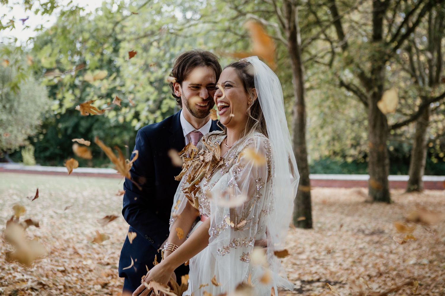 A couple in wedding attire shares joyful moments together while autumn leaves fall around them in a park setting.