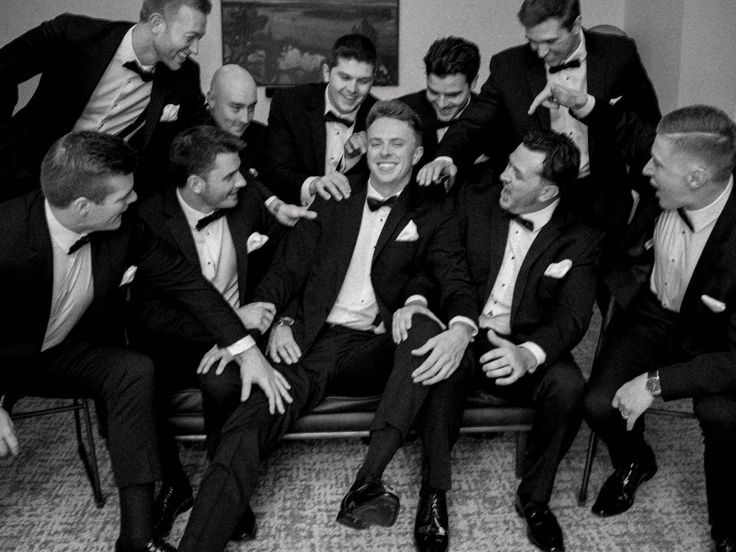 Groomsmen in black suits laughing together in candid black and white photo.
