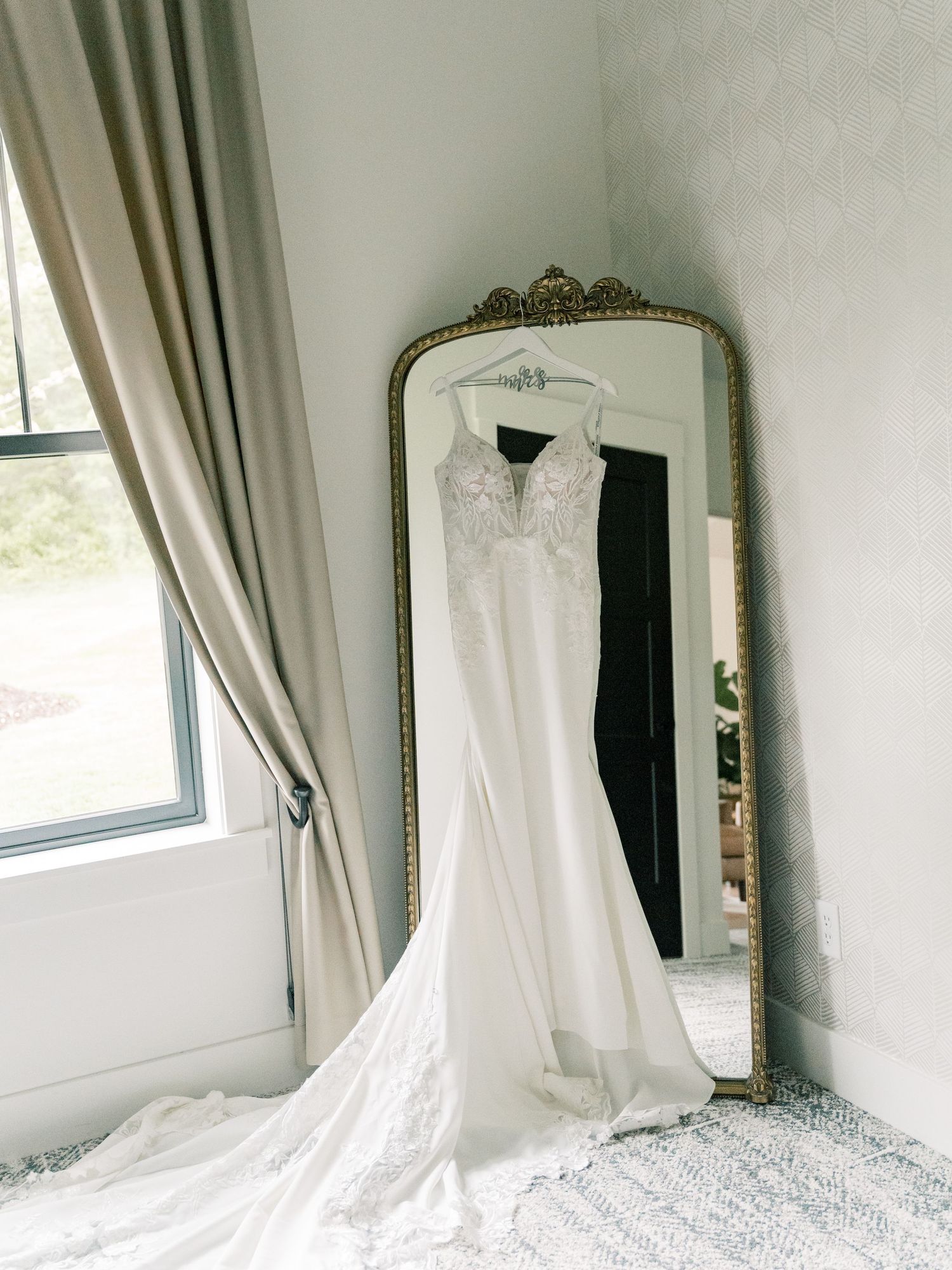 Wedding dress hanging elegantly against white wall with ornate gold mirror and natural window light.