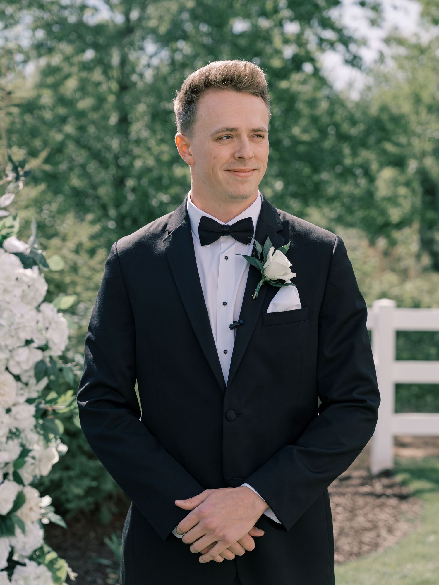 A groom in a classic black tuxedo and white boutonniere poses outdoors for wedding portraits.