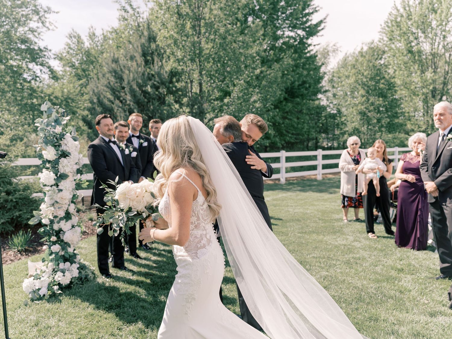 Wedding ceremony guests stand in rows on a lush green lawn with white chairs.