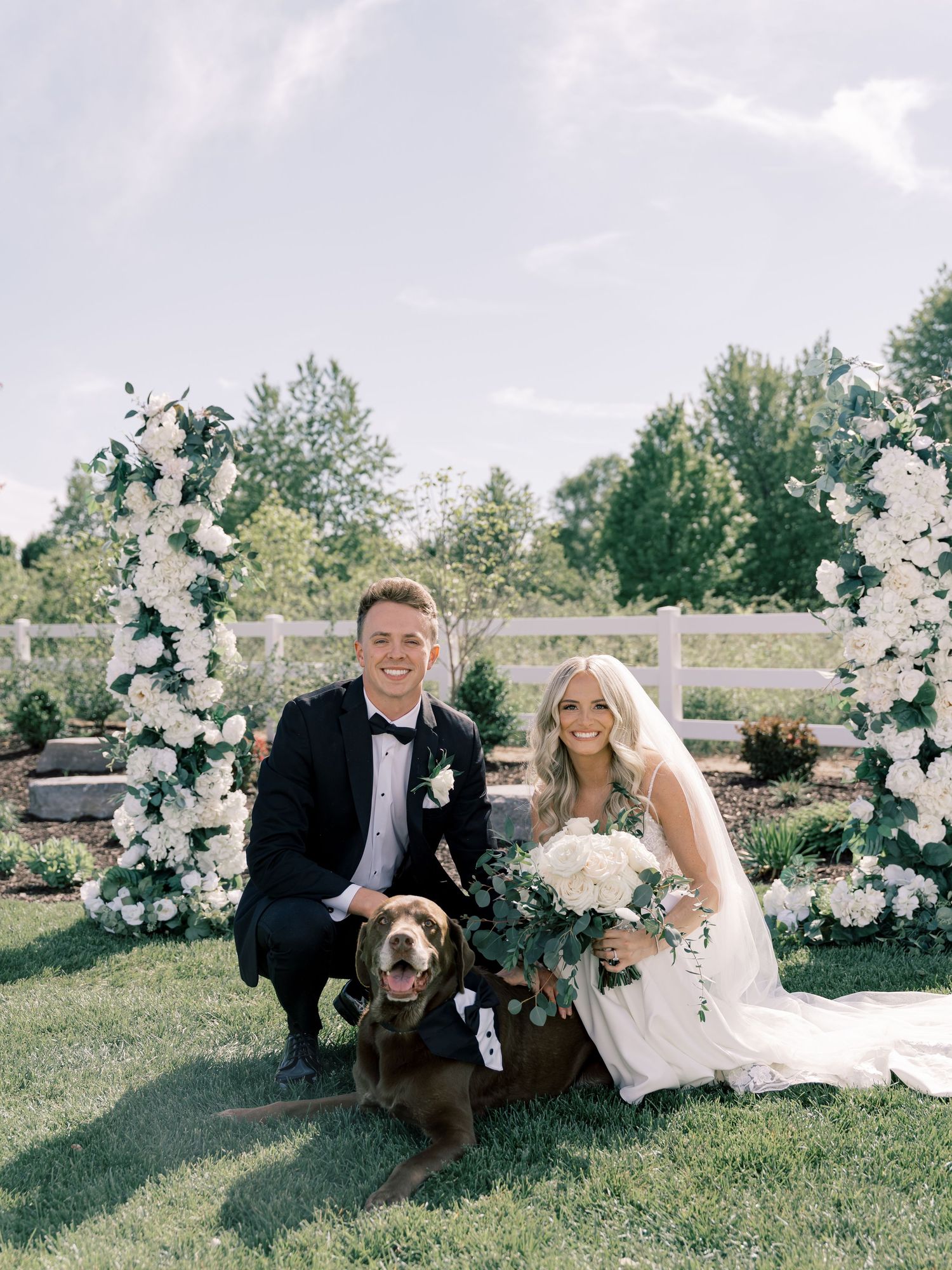 A wedding couple poses with their brown dog on grass near white floral arrangements.