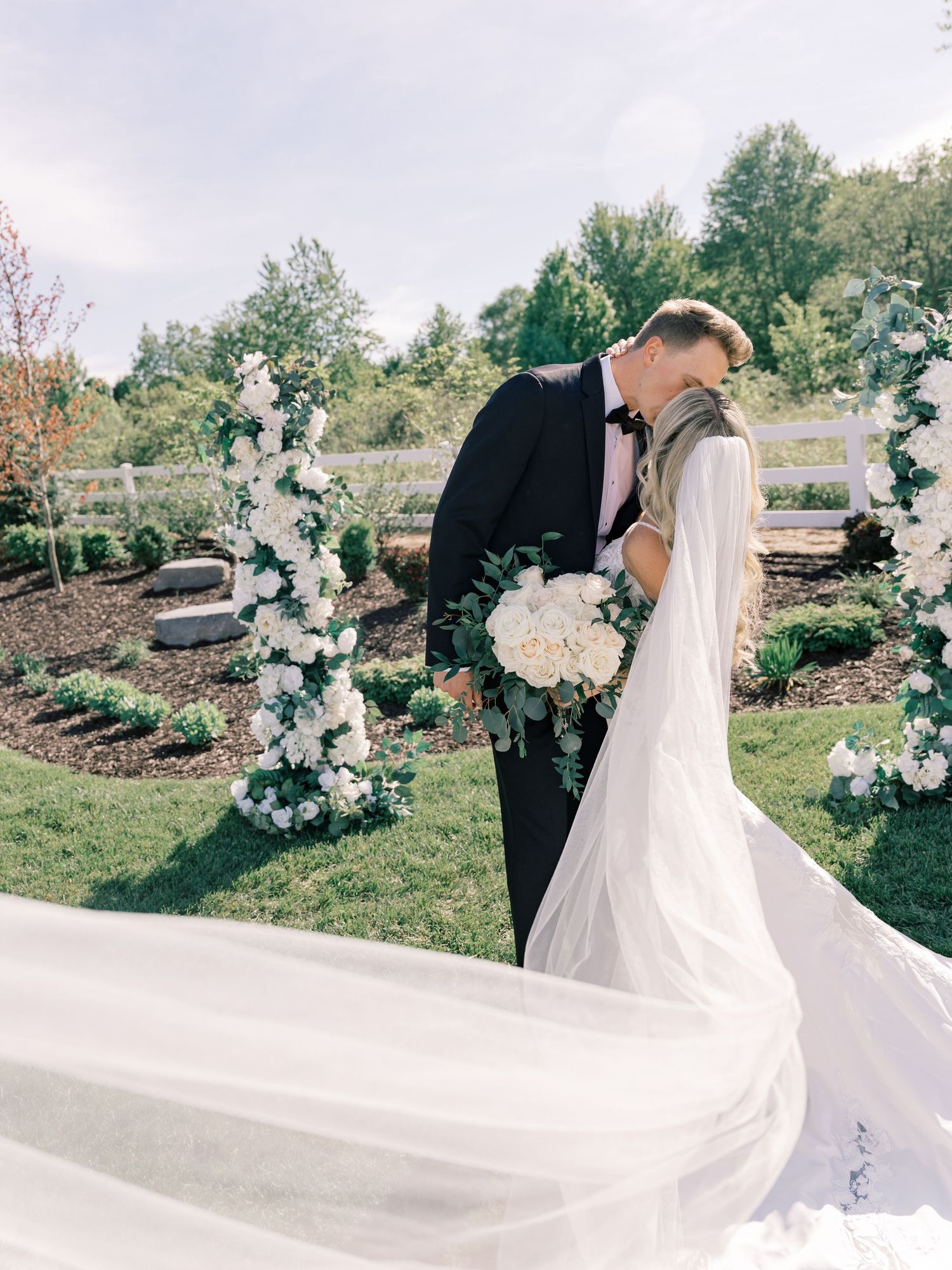 Romantic wedding portrait with flowing white dress veil against an outdoor garden backdrop.