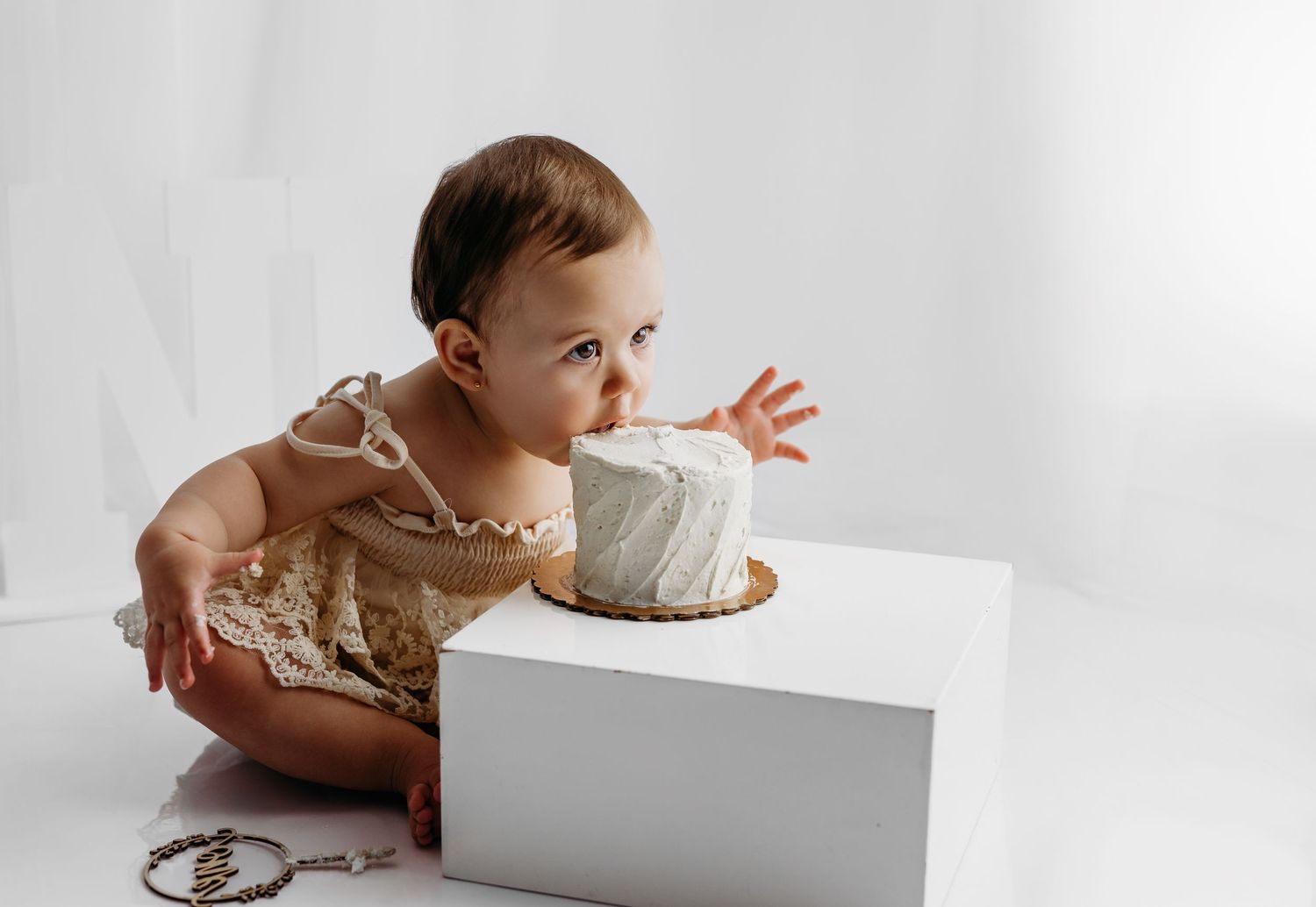 Baby in white lace dress and bow sits on white wooden chair against light backdrop for professional photo shoot.