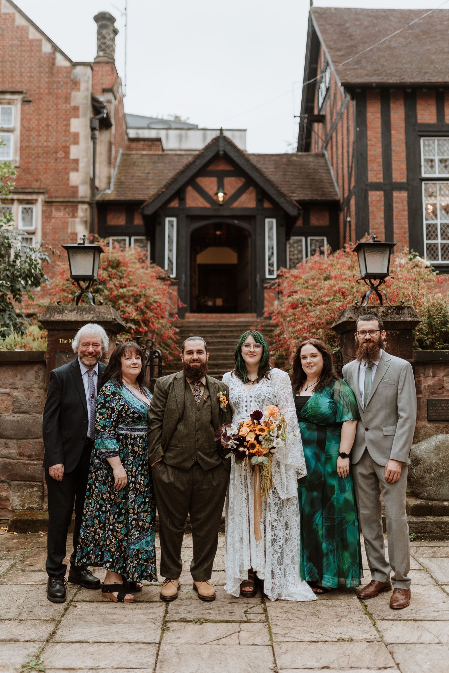 A group of wedding guests pose together outside a brick building with Tudor-style architecture and autumn decor