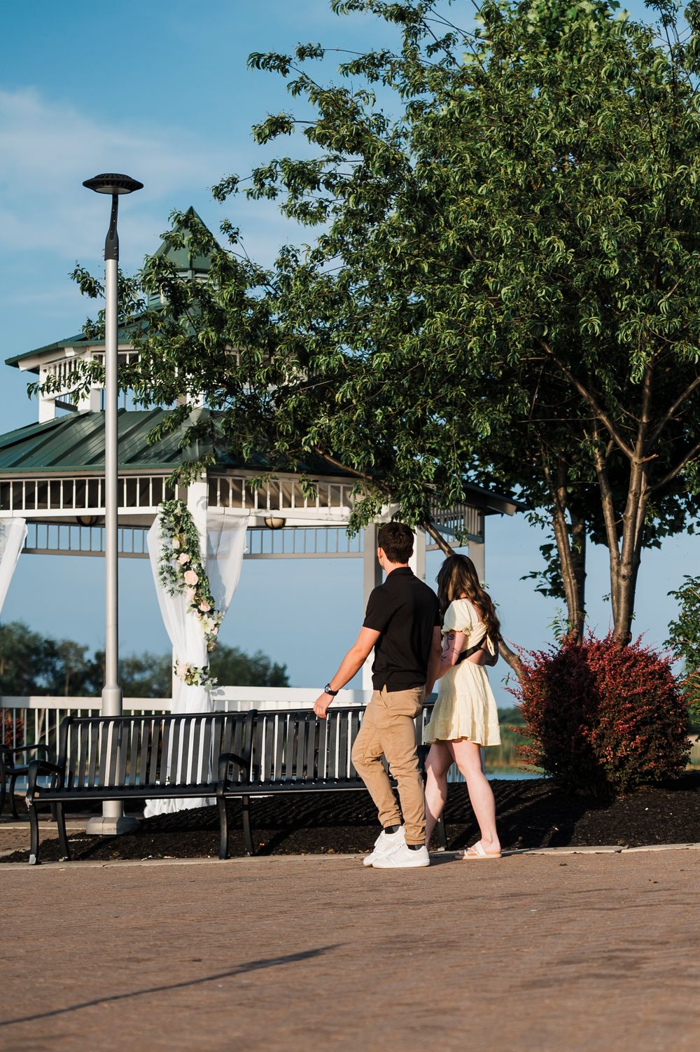 A couple sits together on a brick patio overlooking a scenic view with white gazebo and trees in the background.