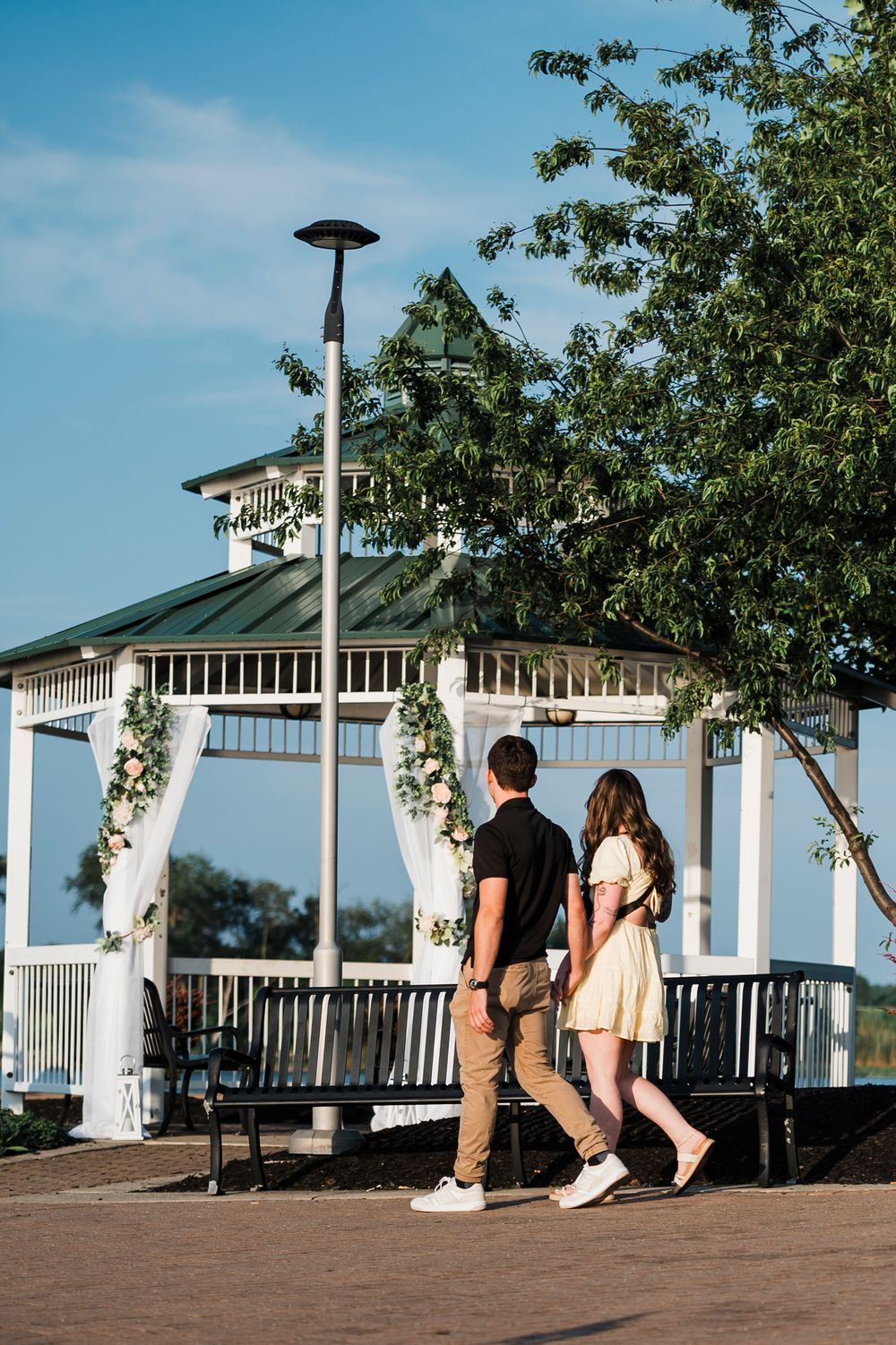 A couple walks past a white gazebo decorated with floral garlands on a sunny summer day.