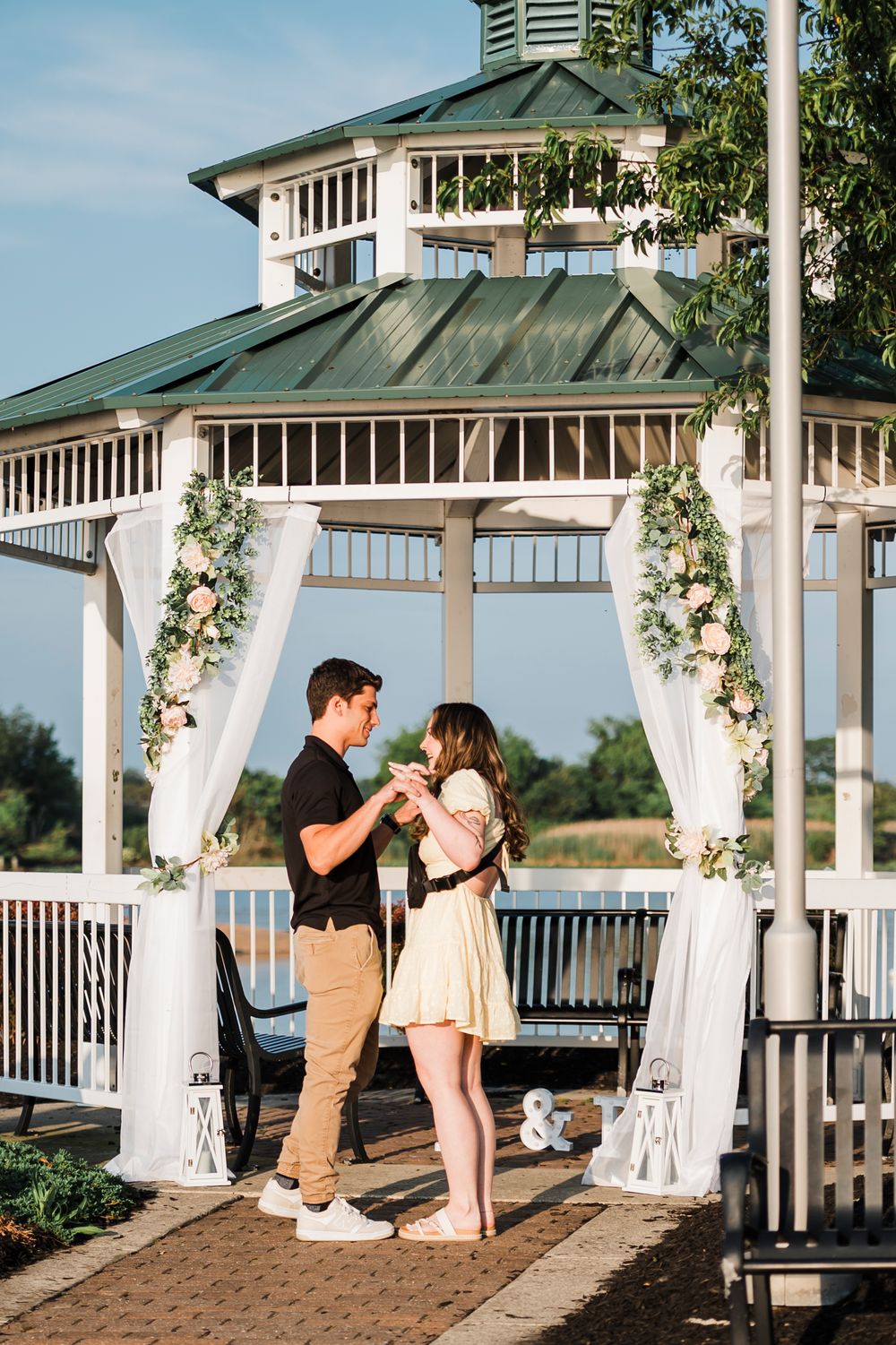 A romantic proposal scene unfolds in a white gazebo decorated with floral arrangements and flowing curtains by the waterfront.